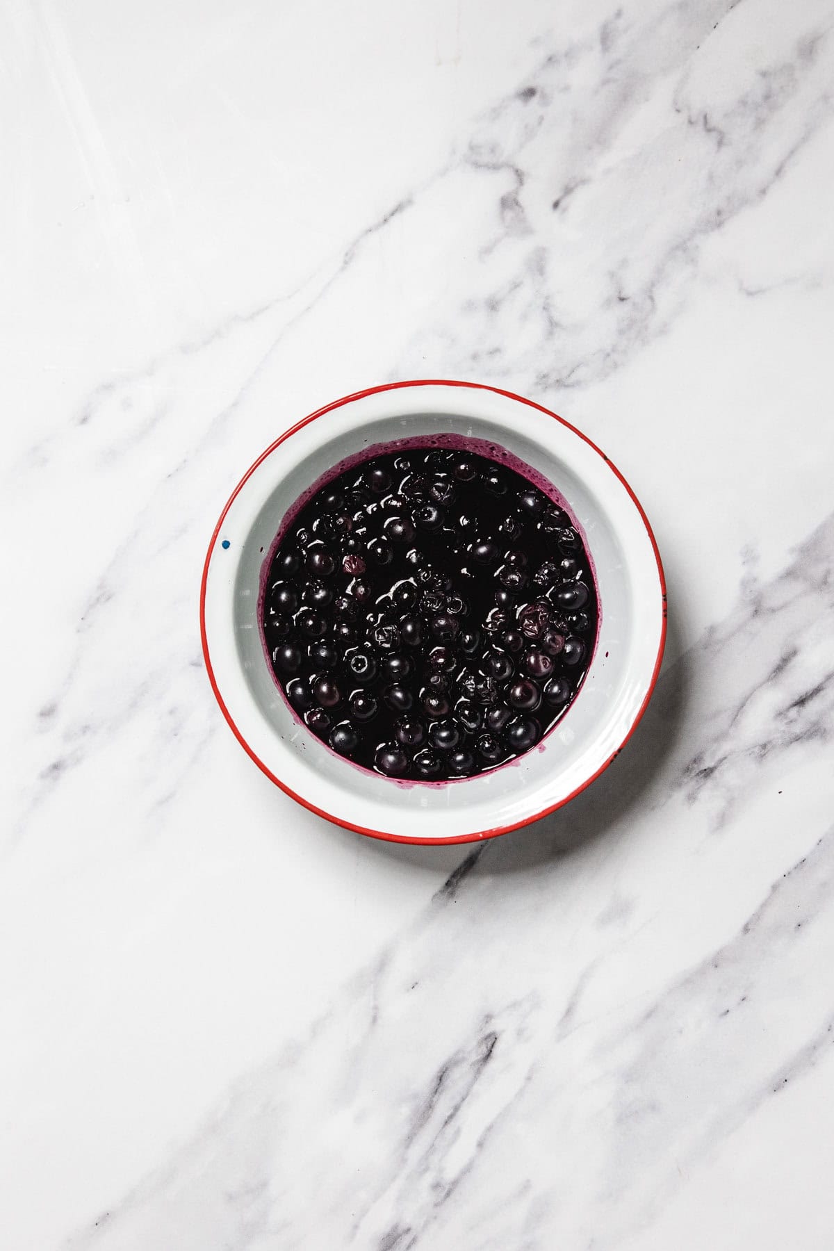 A white bowl with a red rim contains dark purple blueberries in blueberry sauce, placed on a white marble surface with gray veining.