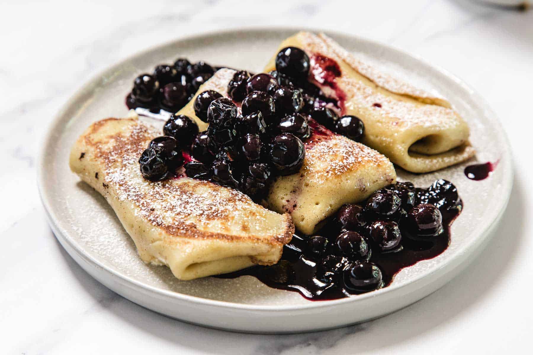 A plate with three folded crepes dusted with powdered sugar, topped with a generous serving of blueberries and rich blueberry sauce on a white surface.