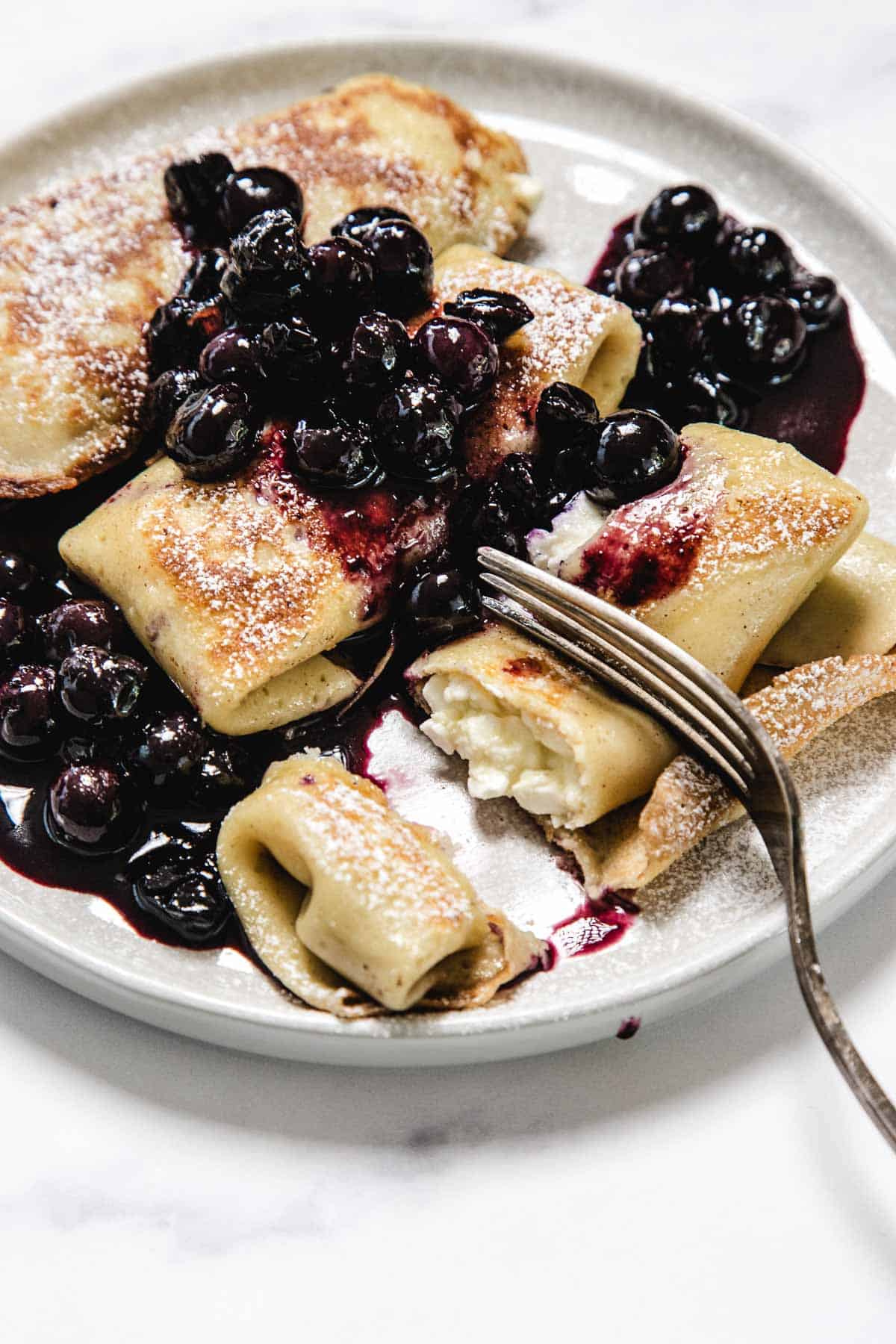 A plate of rolled crepes filled with white cheese, generously topped with blueberry sauce and cooked blueberries, lightly dusted with powdered sugar. A fork rests on the plate, cutting into one of the delicious crepes.