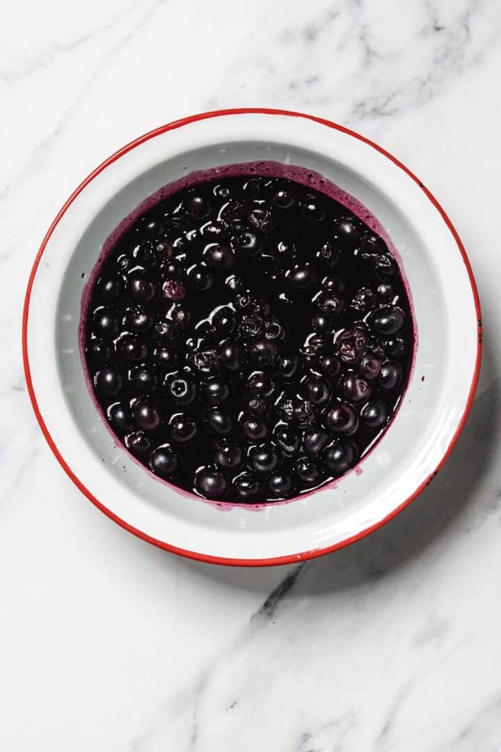 A white bowl with a red rim filled with blueberries in rich Blueberry Sauce, placed on a white marble surface.
