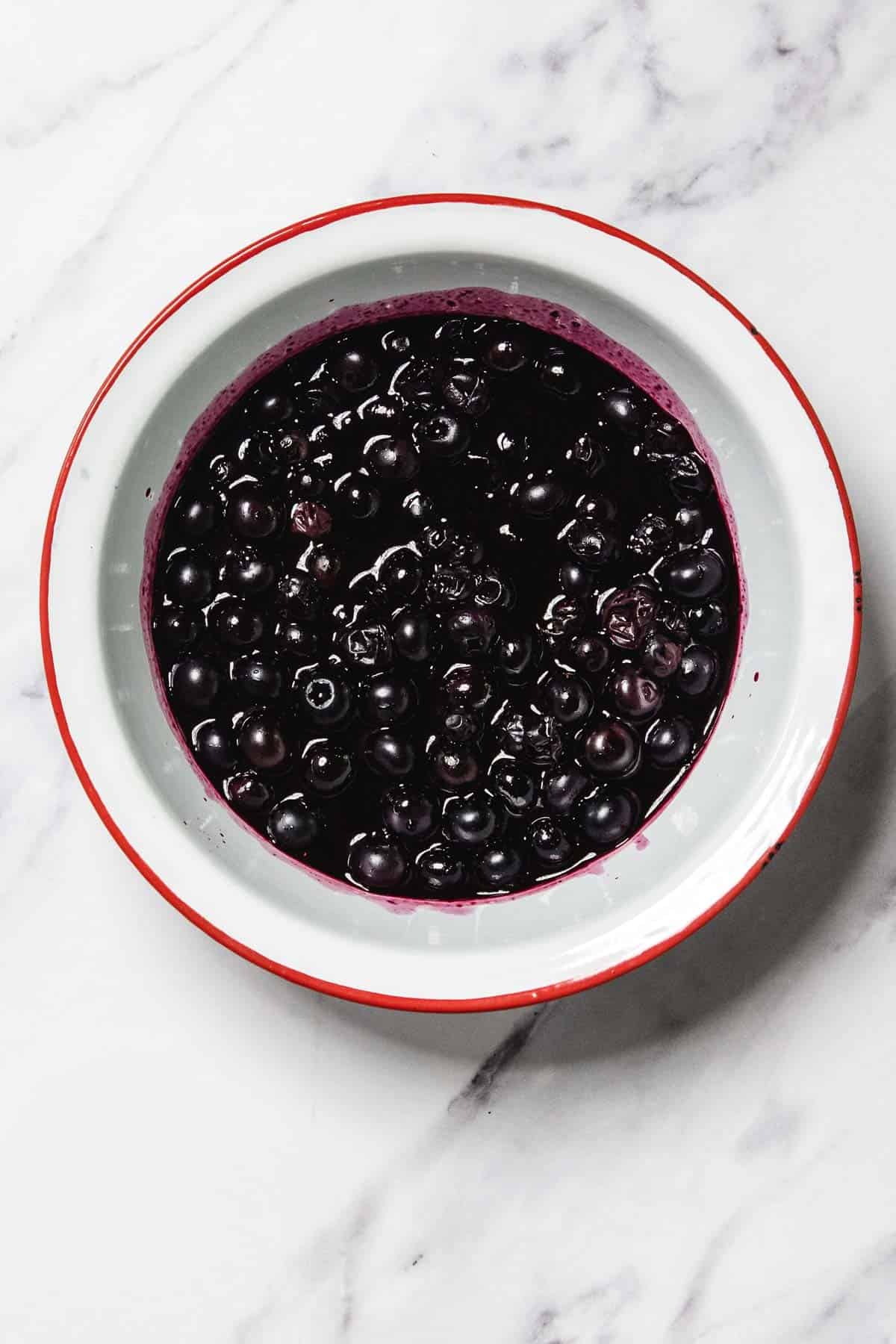 A white bowl with a red rim filled with blueberries in rich Blueberry Sauce, placed on a white marble surface.