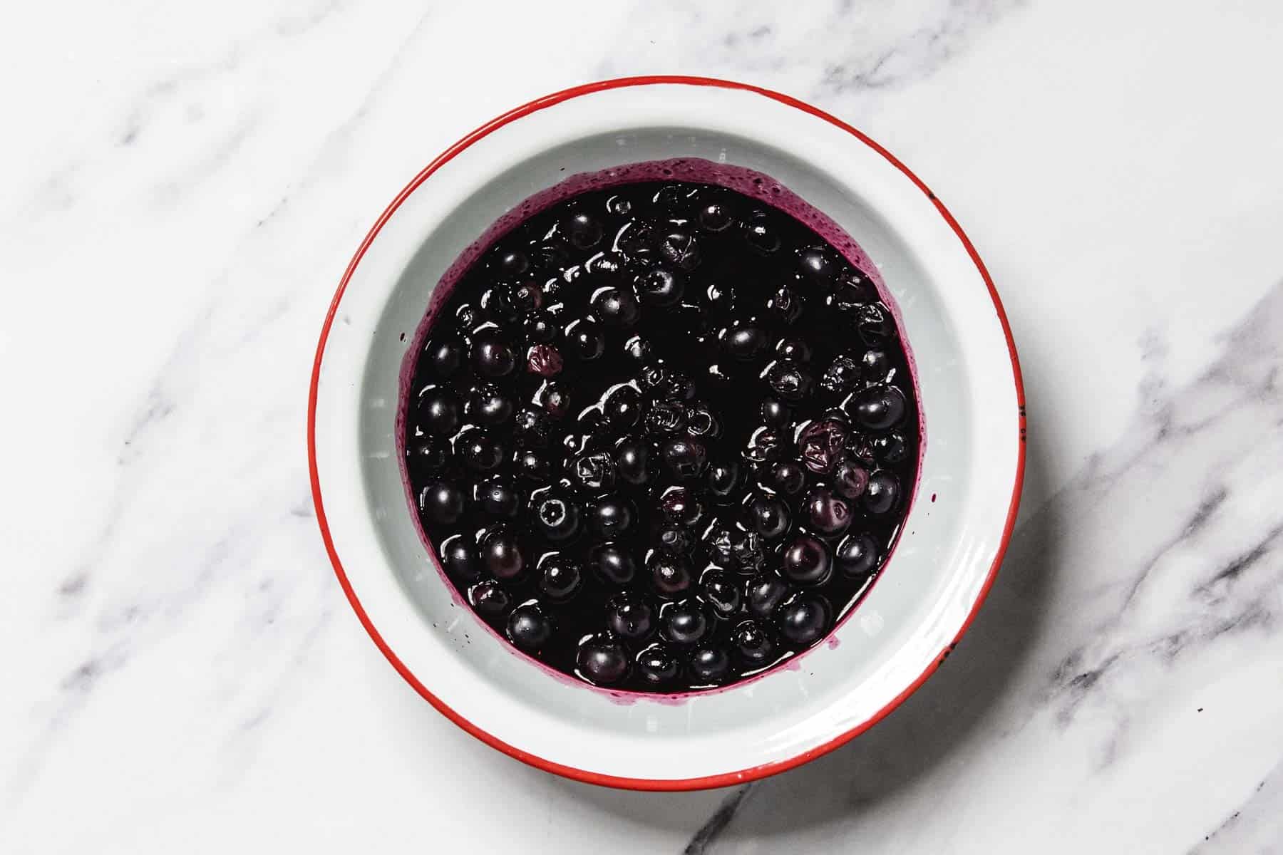 A white bowl with a red rim contains homemade blueberry sauce with stewed blueberries in purple liquid, placed on a white marble surface.