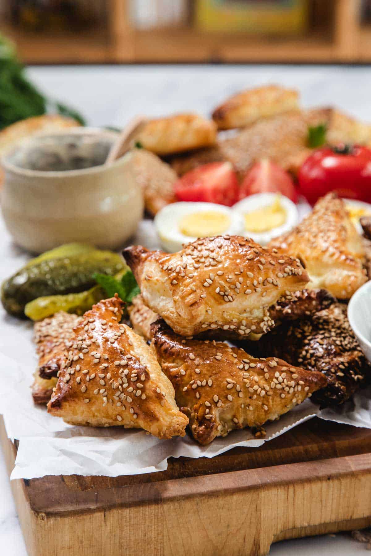 A wooden board with golden, sesame-topped Bourekas triangles, hard-boiled egg slices, pickles, tomatoes, and a small bowl with a spoon—classic Pinukim style—beautifully arranged on parchment paper.