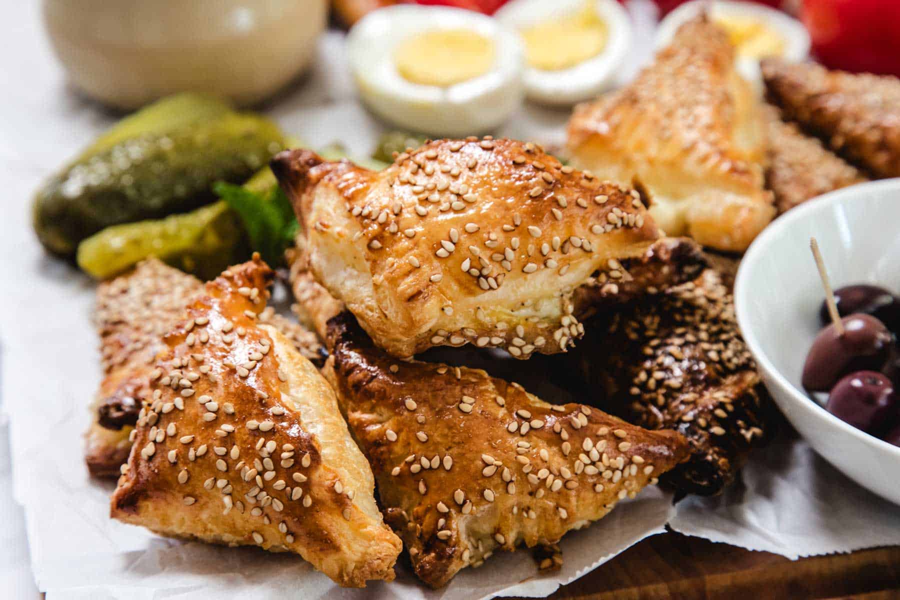 A close-up of flaky, golden-brown Bourekas topped with sesame seeds, stacked on parchment paper, with pickles, olives, and halved boiled eggs visible in the background—a savory Pinukim delight.