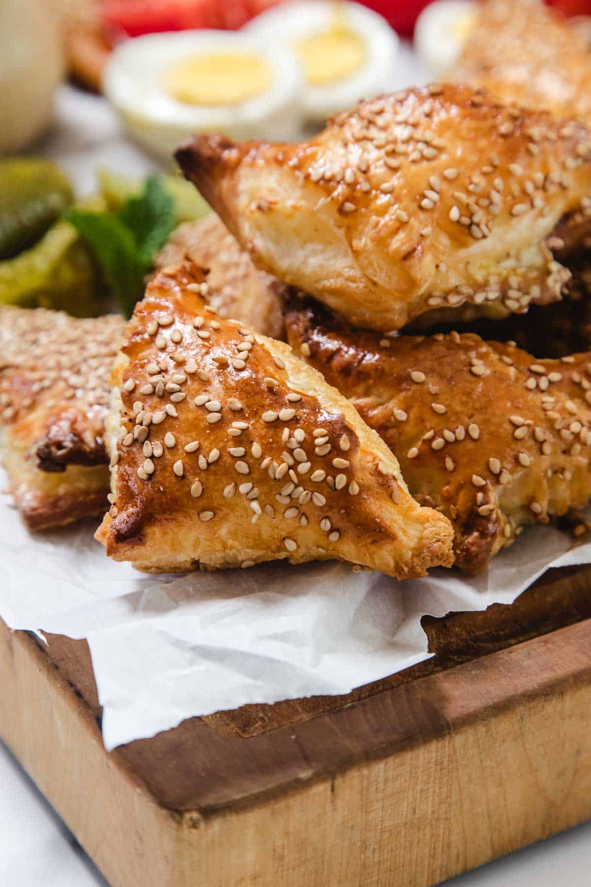 Bourekas Pinukim, classic Israeli pastries sprinkled with sesame seeds, are stacked on parchment paper atop a wooden board, with sliced boiled eggs and pickles visible in the blurred background.
