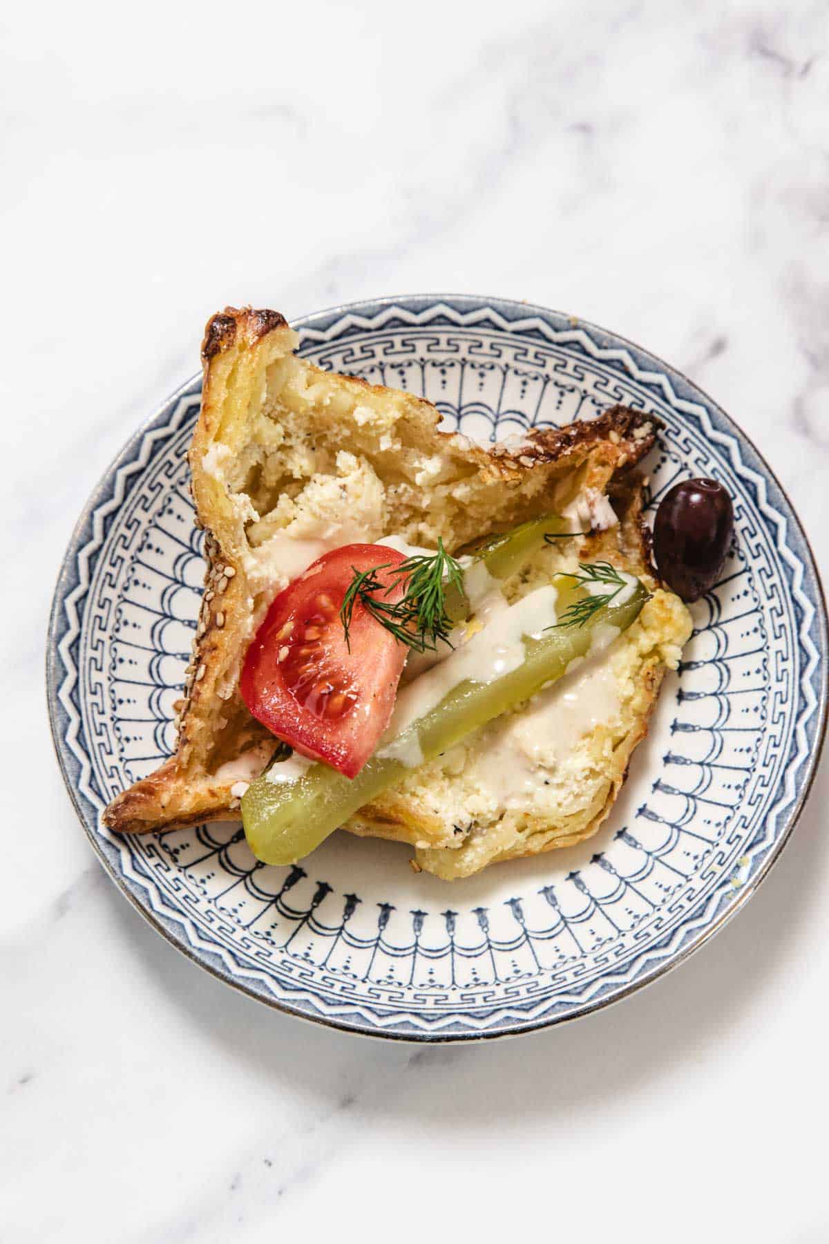 A slice of flaky bourekas bread topped with a tomato wedge, a pickle spear, creamy sauce, fresh dill, and a black olive—these pinukim are served on a patterned blue and white plate set on a white marble surface.