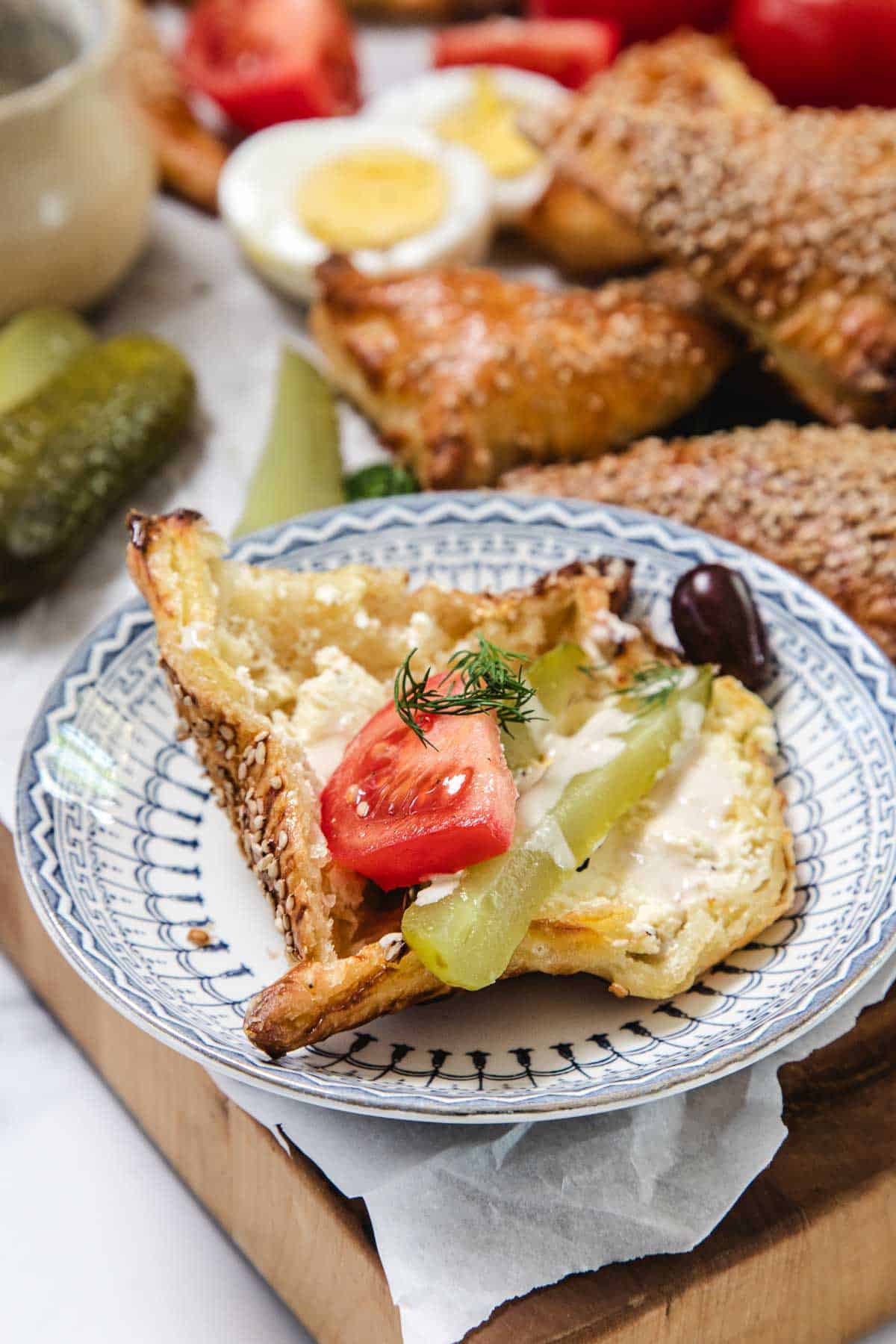 A slice of Bourekas pastry on a patterned plate, topped with tomato, pickle, feta cheese, a sprig of dill, and an olive. Hard-boiled egg halves and more Pinukim pastries are visible in the background.