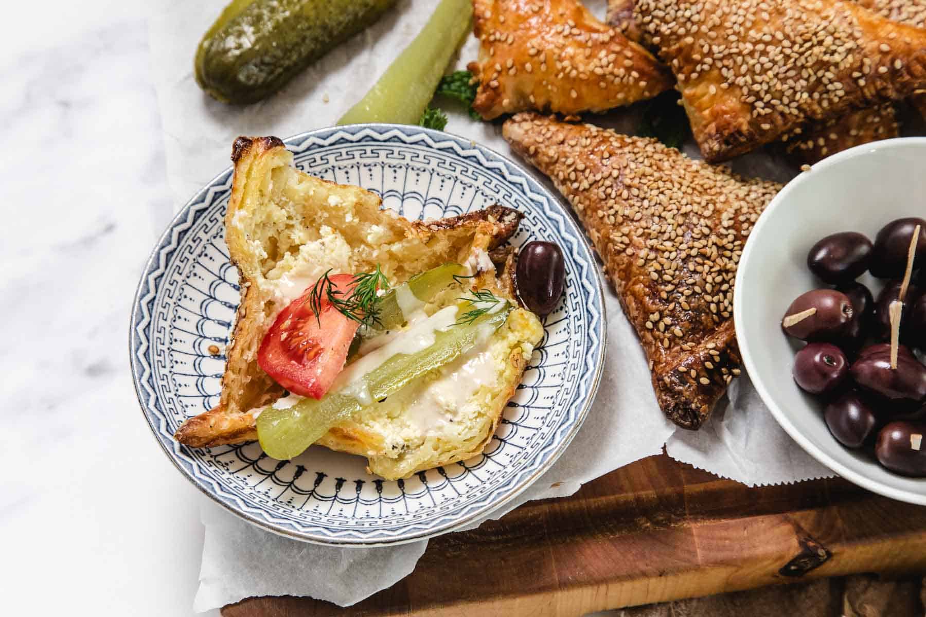 A plate with a toasted sandwich topped with tomato, pickle, and dill sits next to black olives and flaky bourekas on a wooden board. A whole pickle is visible in the background, adding extra pinukim to the scene.