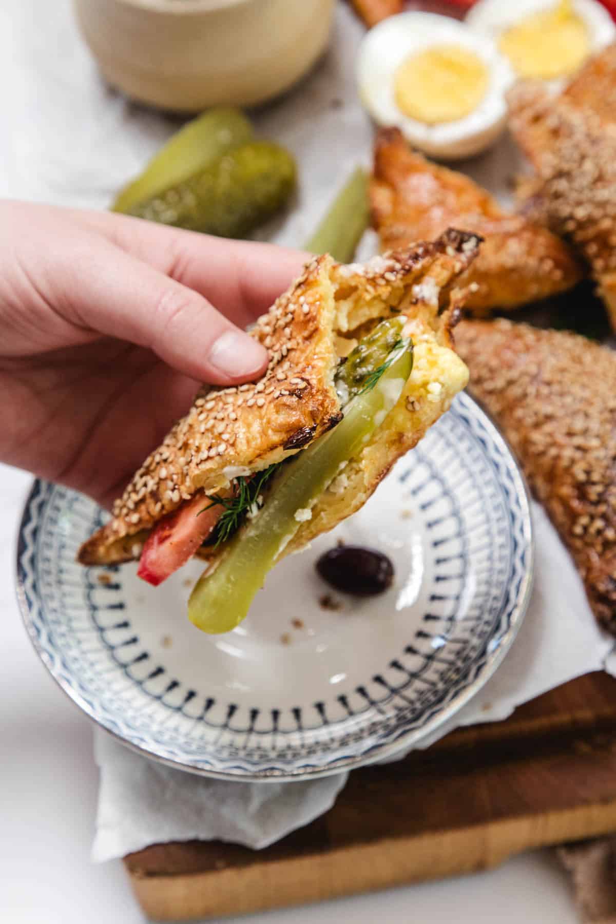 A hand holds a Bourekas pastry filled with egg, pickles, tomato, and herbs over a patterned plate. In the background, more pastries—perfect Pinukim—join a hard-boiled egg and pickles on a wooden board.