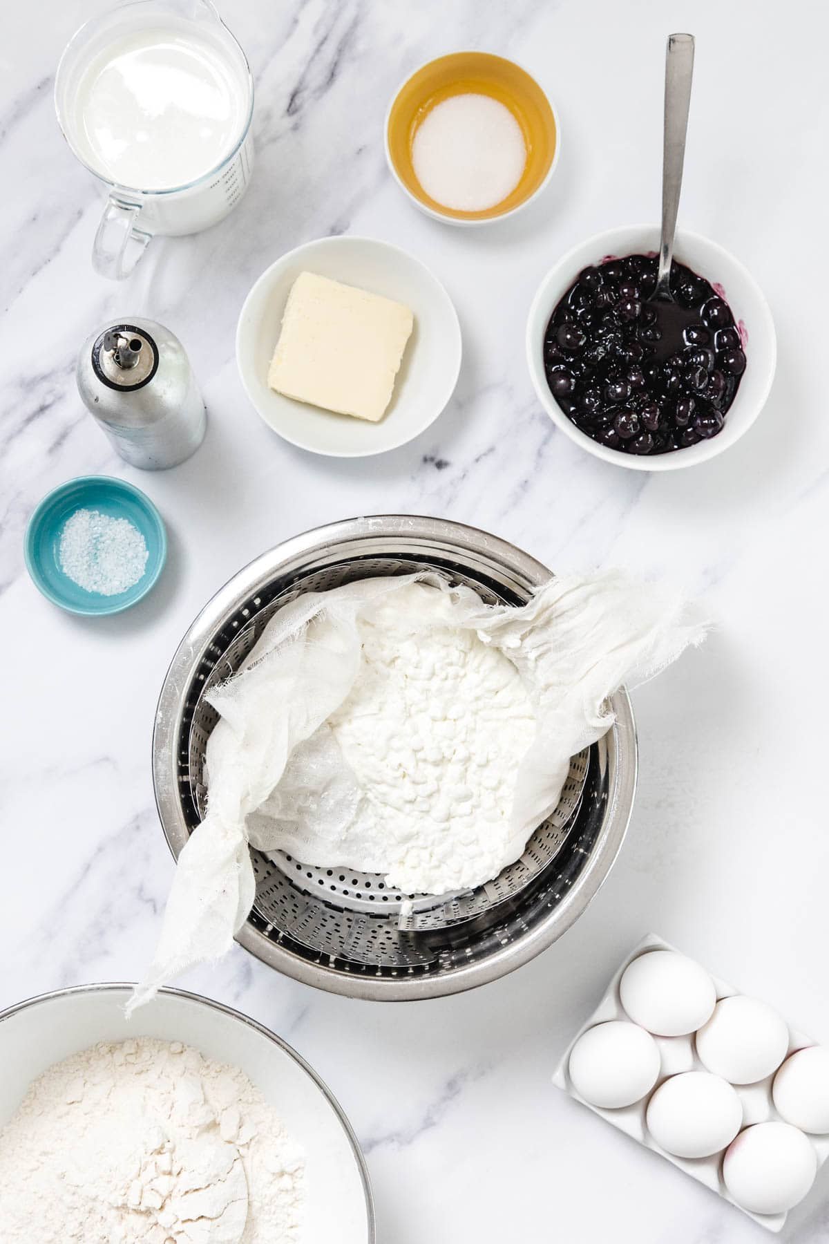 A marble countertop displays ingredients for Cheese Blintzes, including a bowl of flour, eggs in a carton, sea salt, coarse salt, butter, sugar, milk, blueberry compote, and ricotta cheese draining in cheesecloth over a colander.
