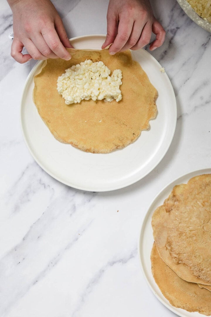 A person folds a thin pancake on a white plate, placing a cheese blintzes filling of crumbled white cheese inside. Another plate with stacked pancakes sits nearby on a marble surface.