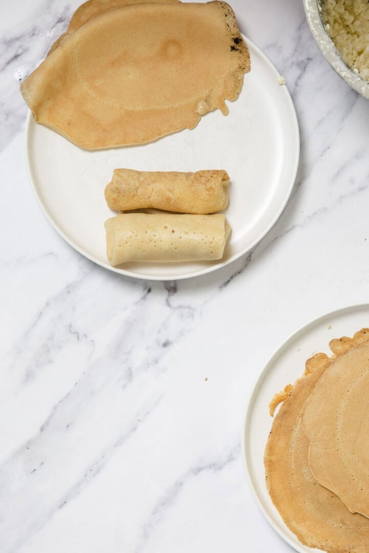 Two white plates with thin, round crepes. One plate has two crepes rolled up like cheese blintzes and an unrolled crepe above them. The other plate holds a stack of whole crepes on a white marble surface.