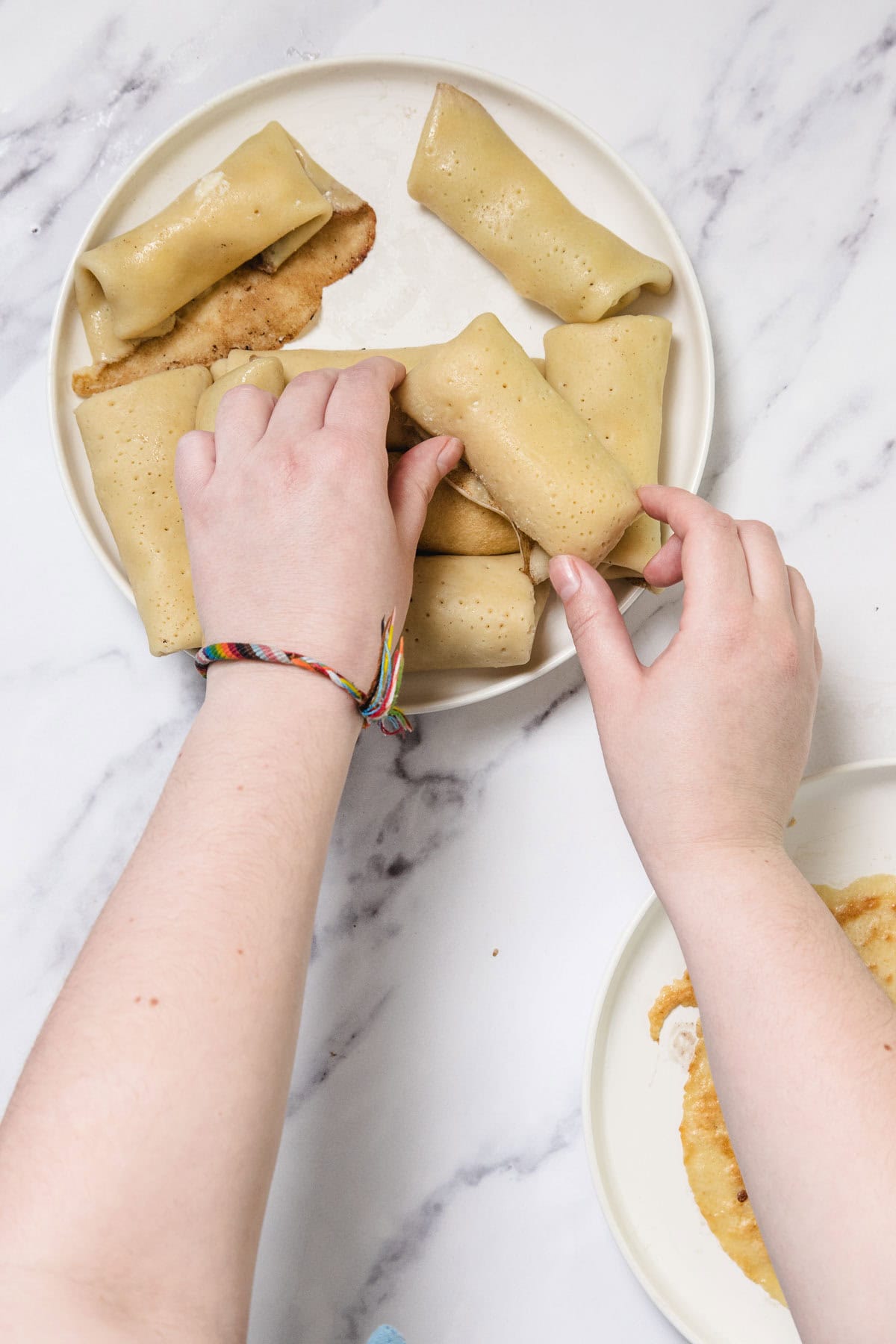 A pair of hands places rolled Cheese Blintzes onto a white plate with more crepes and a fried item visible; the person is wearing a colorful braided bracelet, and the background is a white marble surface.