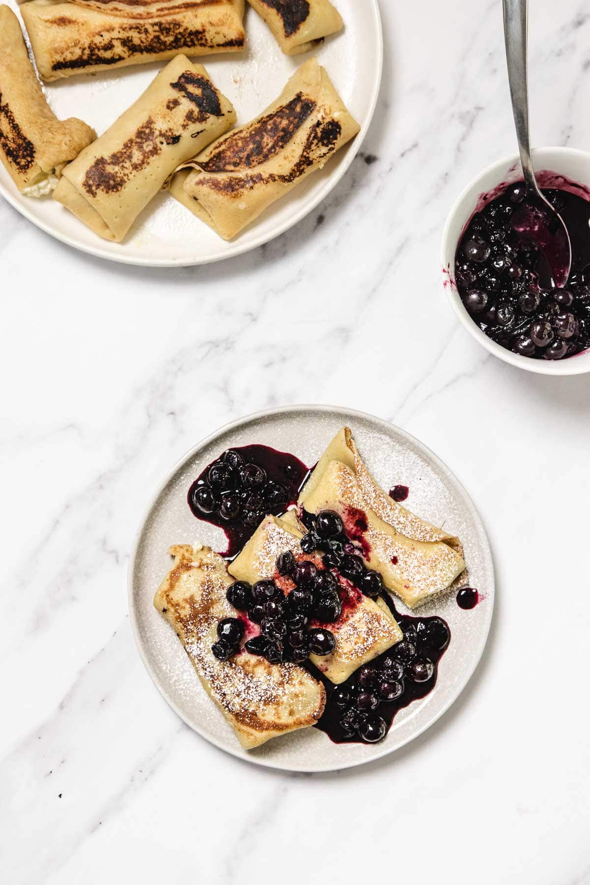 A plate with three rolled crepes topped with blueberry sauce and powdered sugar sits beside a bowl of extra blueberry sauce and another plate with more crepes on a white marble surface.
