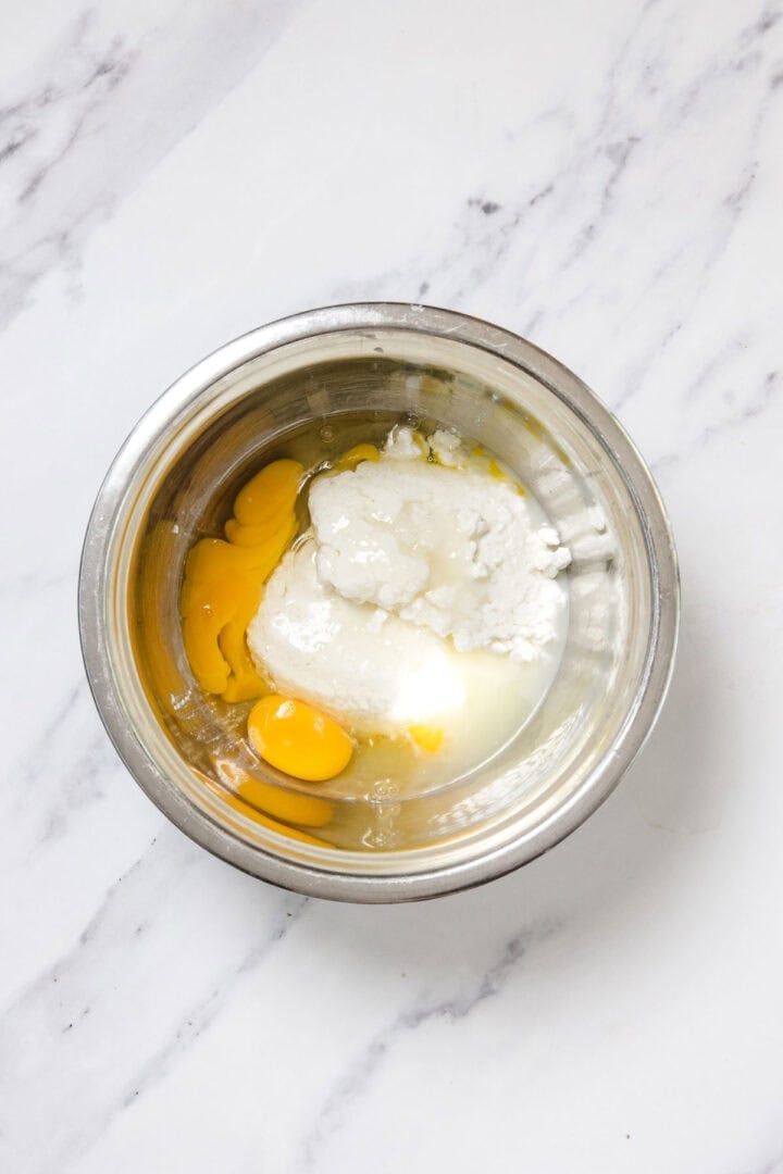 A metal mixing bowl on a white marble countertop contains eggs, yogurt, and flour—ingredients coming together for delicious Cheese Blintzes before being mixed.