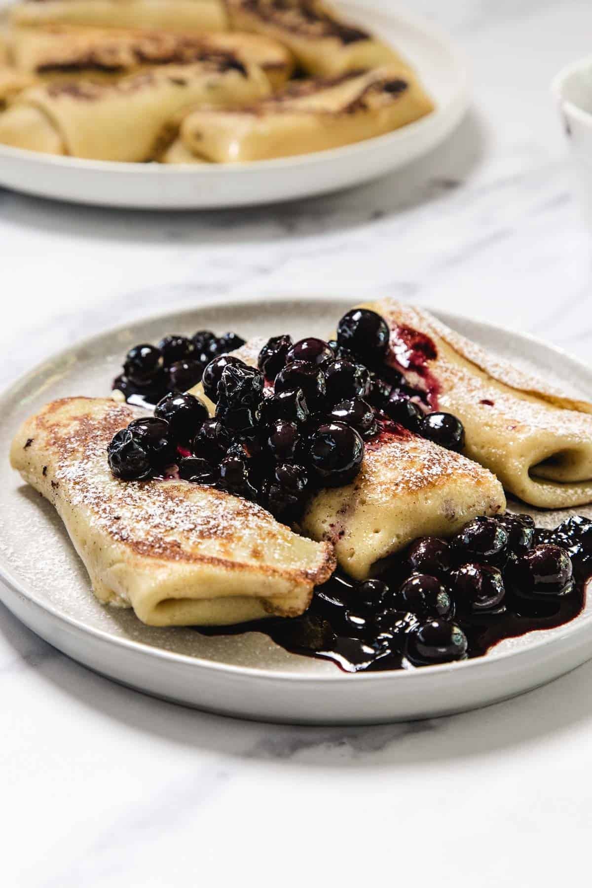 Three rolled Cheese Blintzes on a gray plate are topped with a blueberry compote. A second plate with more blintzes is visible in the background on a white surface.