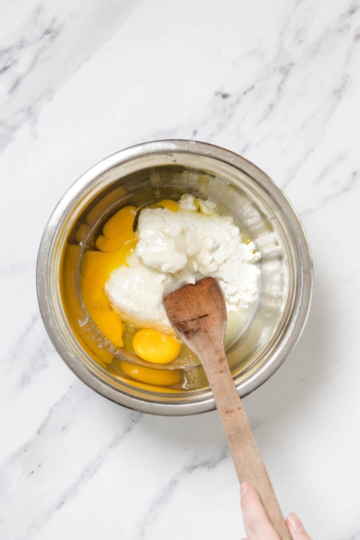 A metal mixing bowl containing eggs and a white creamy mixture for cheese blintzes sits on a marble surface. A wooden spoon rests inside the bowl, and a hand holds the spoon, ready to mix the ingredients.