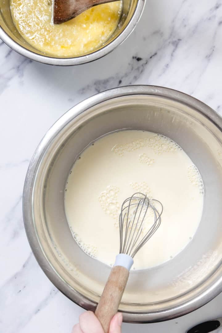 A hand holding a whisk mixes a creamy liquid in a metal bowl on a marble surface—perfect for preparing Cheese Blintzes. Above, another metal bowl holds a yellow mixture with a wooden spoon, ready for the next step.