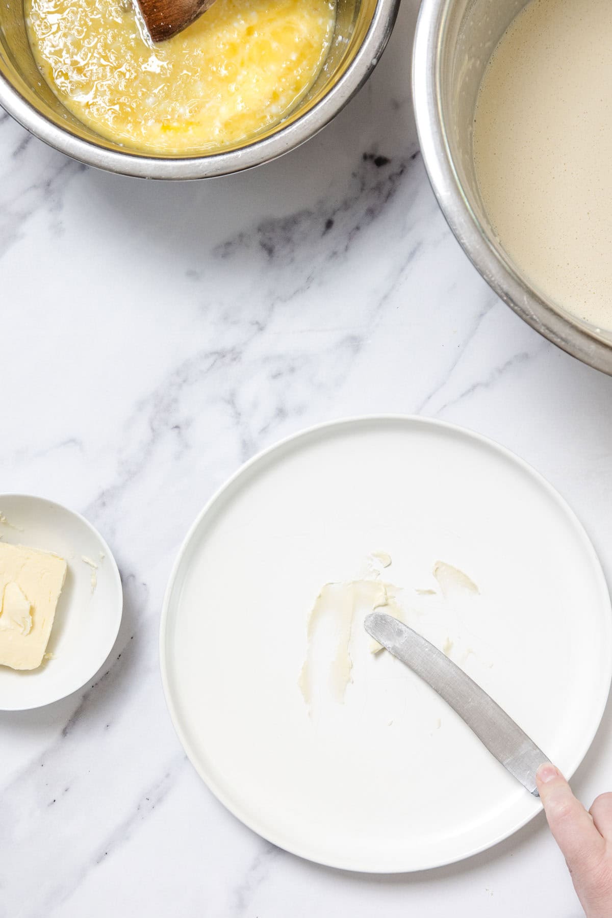 A hand uses a butter knife to spread butter on a white plate. Nearby are ingredients for Cheese Blintzes: a small plate with butter, a bowl of beaten eggs, and a bowl of batter, all set on a marble surface.