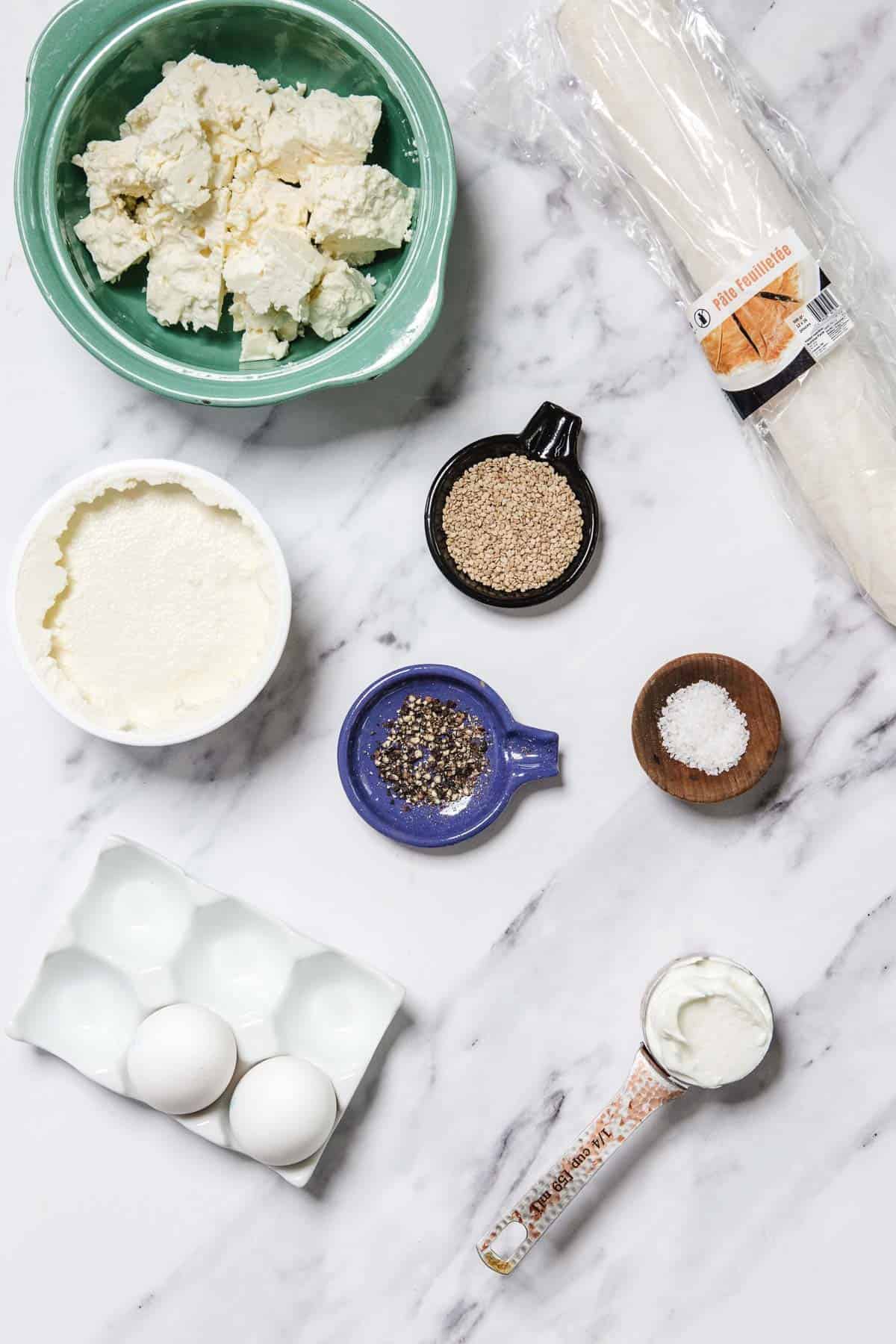 A marble countertop displays ingredients for Bourekas Pinukim: feta in a green bowl, ricotta in white, three eggs, phyllo dough, and small bowls of sesame seeds, black pepper, salt, and a spoonful of yogurt.