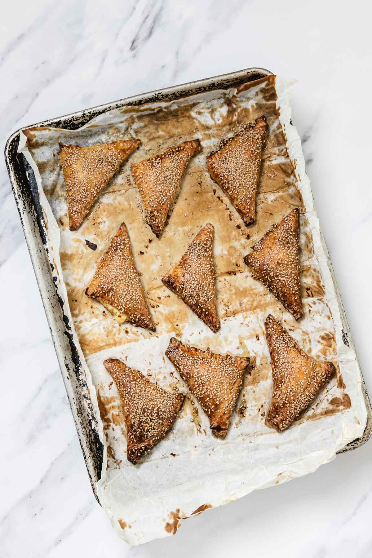 A baking tray lined with parchment paper holds nine golden-brown, triangular Cheese Bourekas topped with sesame seeds. The tray shows signs of use and sits on a white marble surface.