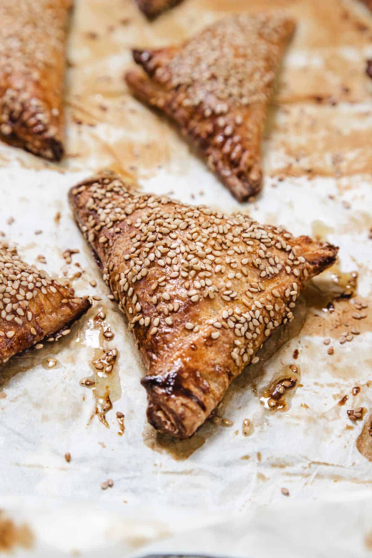 A close-up of Cheese Bourekas, triangular pastries topped with sesame seeds on a sheet of parchment paper. The Cheese Bourekas are browned and appear freshly baked, with visible glaze and some seeds scattered around.