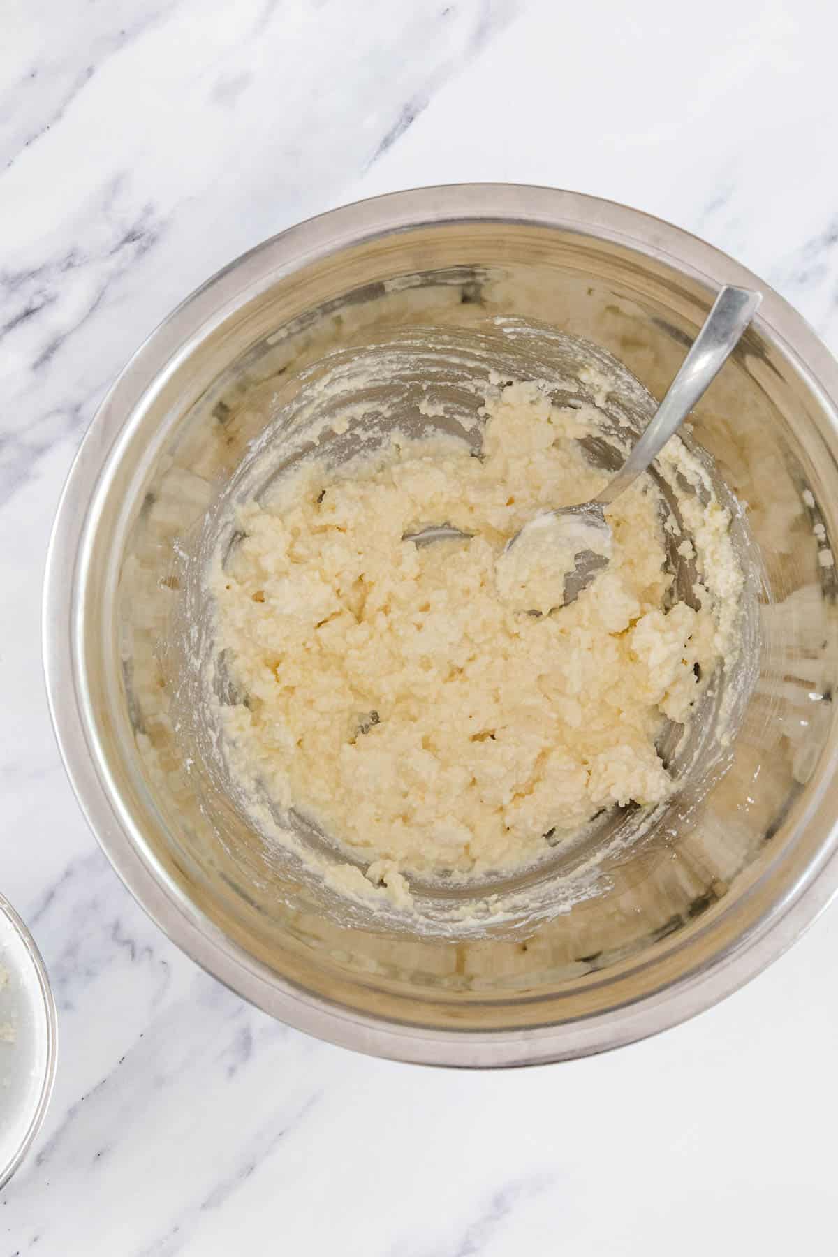 A metal mixing bowl containing a pale, lumpy Cheese Bourekas dough mixture with a metal spoon resting inside, placed on a white marble surface.