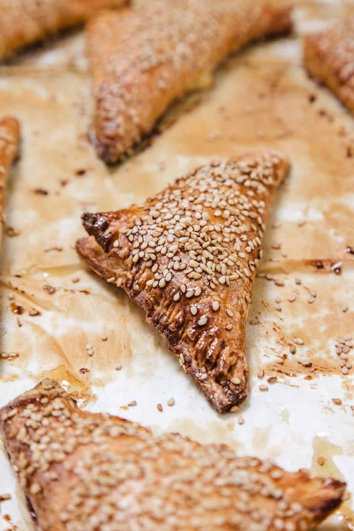 Cheese Bourekas, triangle-shaped pastries topped with sesame seeds, rest on a piece of parchment paper. Their golden-brown, flaky crust is perfectly baked and neatly arranged on the baking surface.