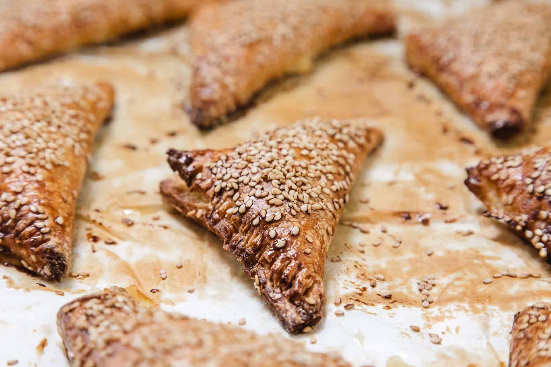 Golden brown Cheese Bourekas, topped with sesame seeds, are arranged on a parchment-lined baking sheet. The triangular pastries appear flaky with a slightly shiny surface and are spaced out on the sheet.