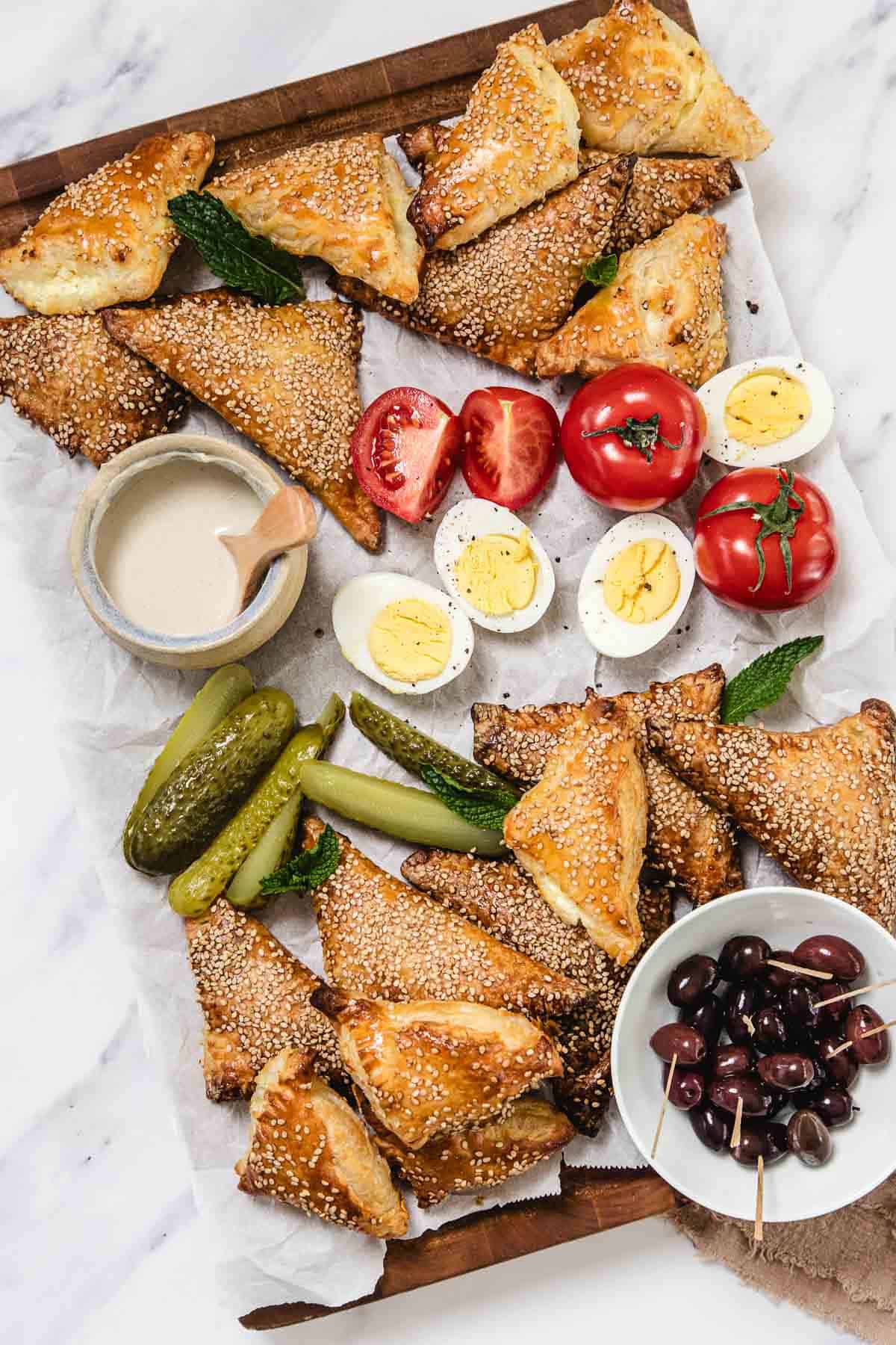 A wooden tray with cheese bourekas topped with sesame seeds, halved hard-boiled eggs, sliced tomatoes, whole tomatoes, pickles, a bowl of olives with toothpicks, and a small bowl of sauce on parchment paper.