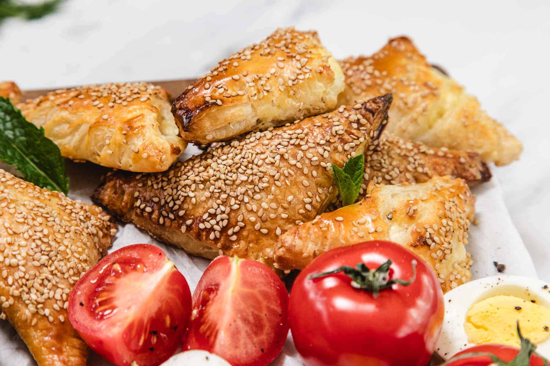A stack of golden Cheese Bourekas topped with sesame seeds sits on parchment paper, accompanied by halved cherry tomatoes, a whole tomato, sliced boiled egg, and fresh mint leaves on a white surface.