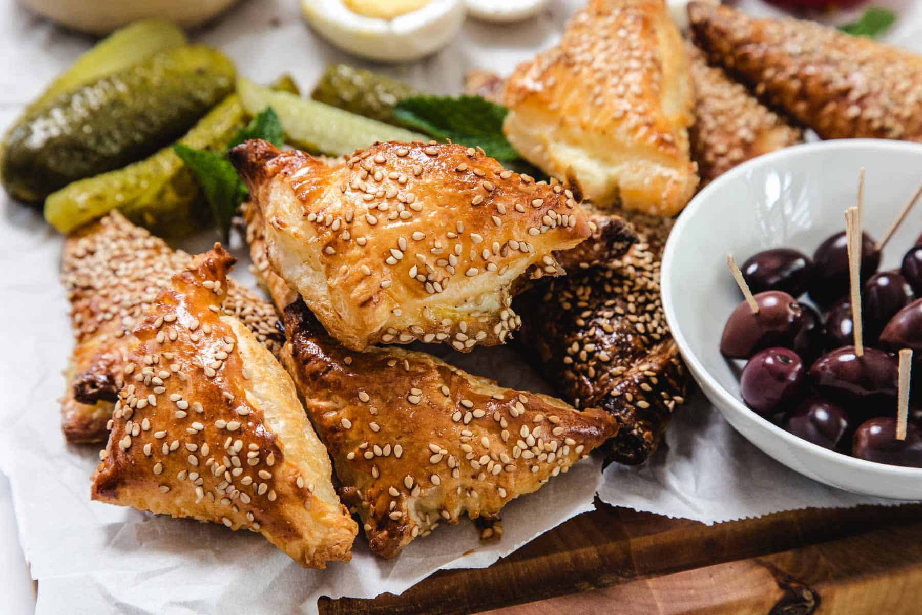 A close-up of golden, triangular Cheese Bourekas topped with sesame seeds on parchment paper, next to sliced pickles and a white bowl filled with black olives on toothpicks.