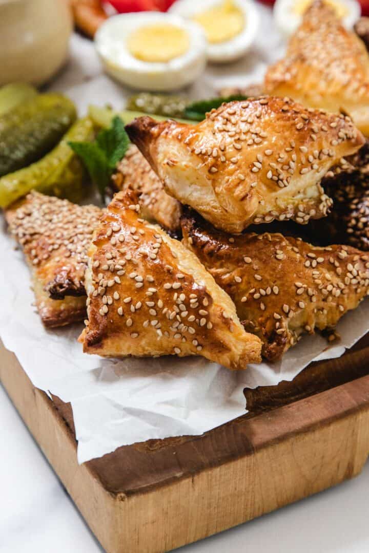 A stack of golden, sesame seed-topped Cheese Bourekas triangles sits on parchment paper, with sliced pickles, mint leaves, and halved boiled eggs visible in the background.