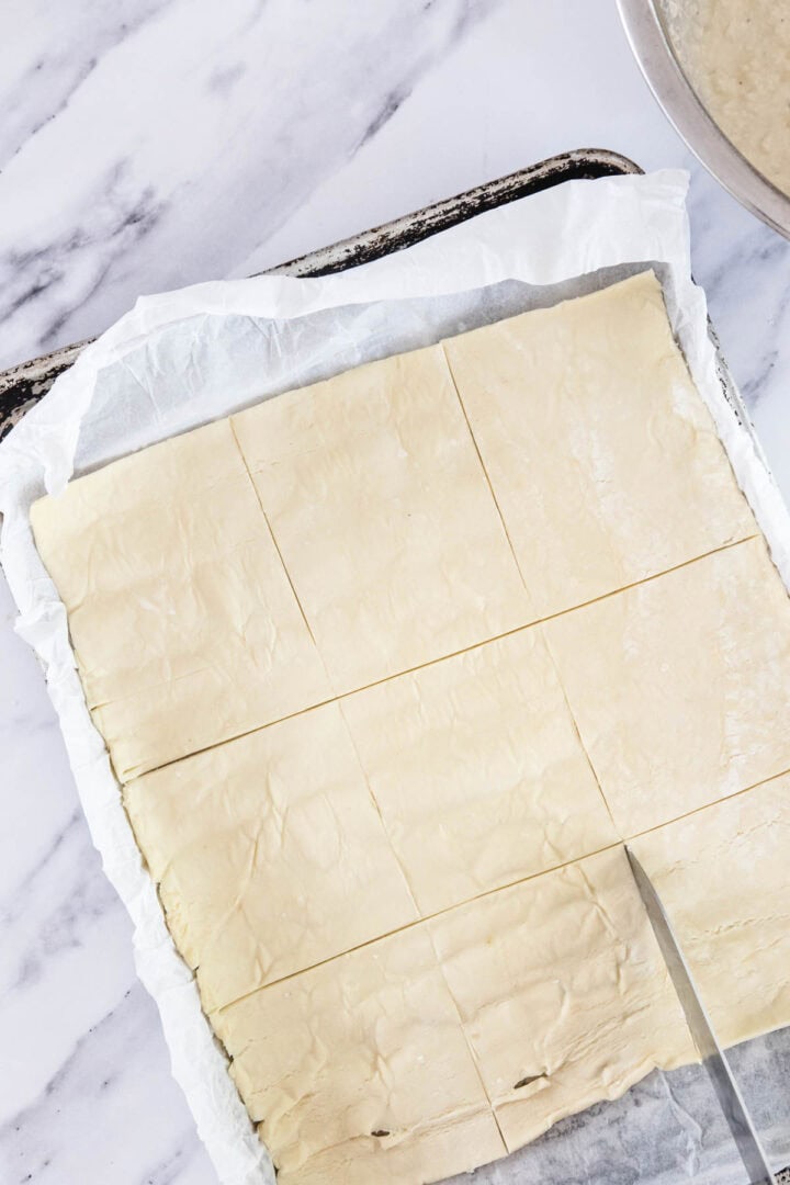 A sheet of puff pastry on parchment paper is cut into nine even rectangles with a knife, ready to be transformed into delicious Cheese Bourekas on a baking tray atop a marble surface.