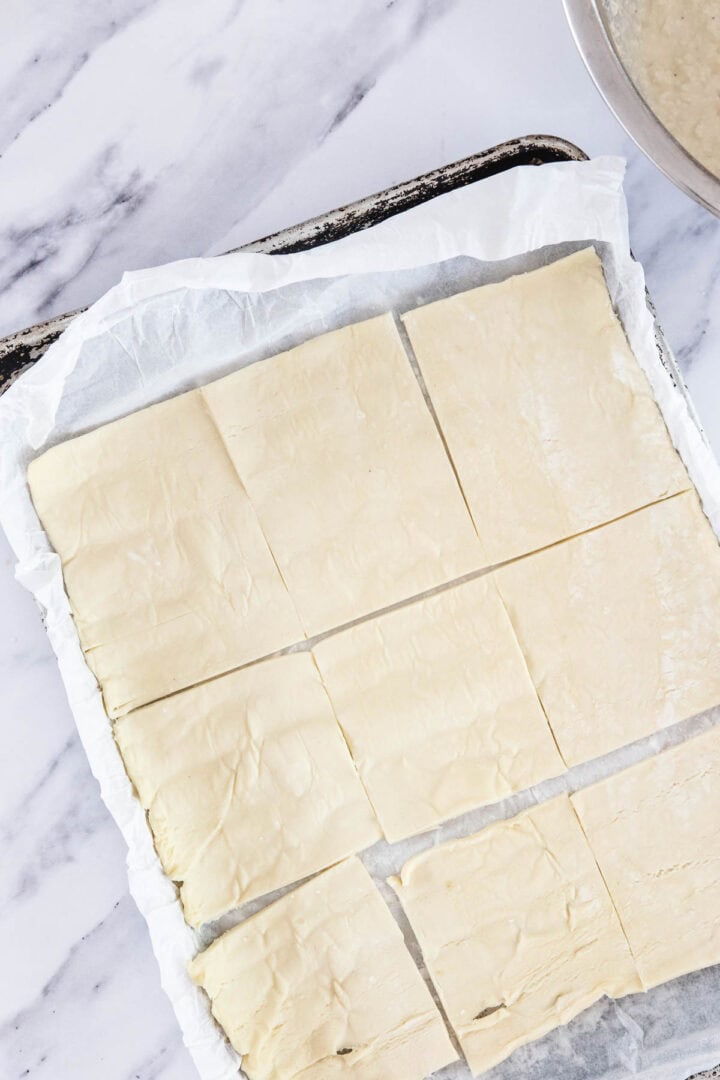 A sheet of puff pastry cut into eight rectangular pieces, ready to become delicious Bourekas Pinukim, rests on parchment paper atop a baking tray on a marble countertop.