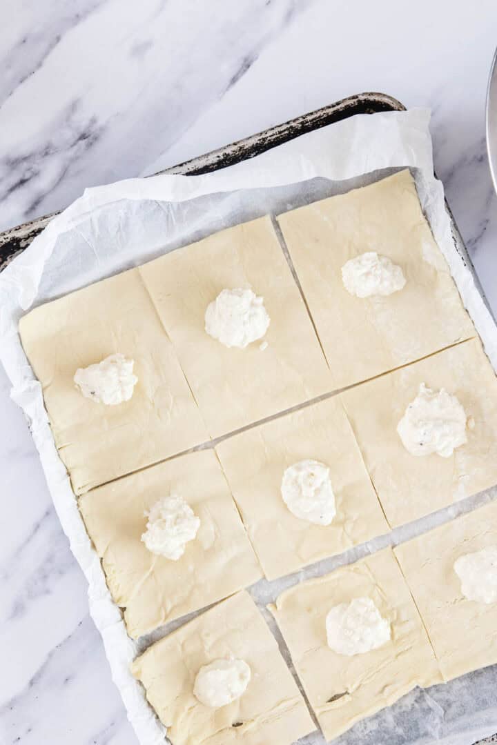 A baking sheet lined with parchment paper holds a sheet of puff pastry cut into nine squares. Each square has a small dollop of white cheese filling in the center, ready to be folded into classic Pinukim bourekas.