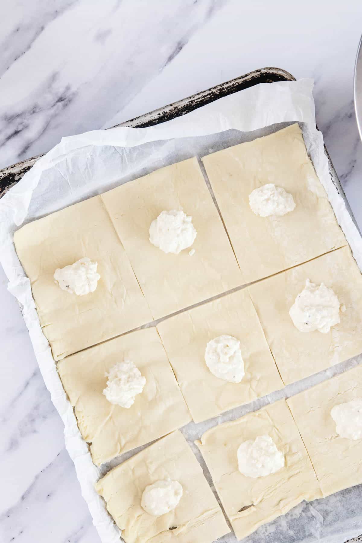 A sheet of puff pastry cut into rectangles on a parchment-lined baking tray, each piece topped with a small dollop of white cheese filling, ready to be baked into delicious Cheese Bourekas, sits on a marble surface.
