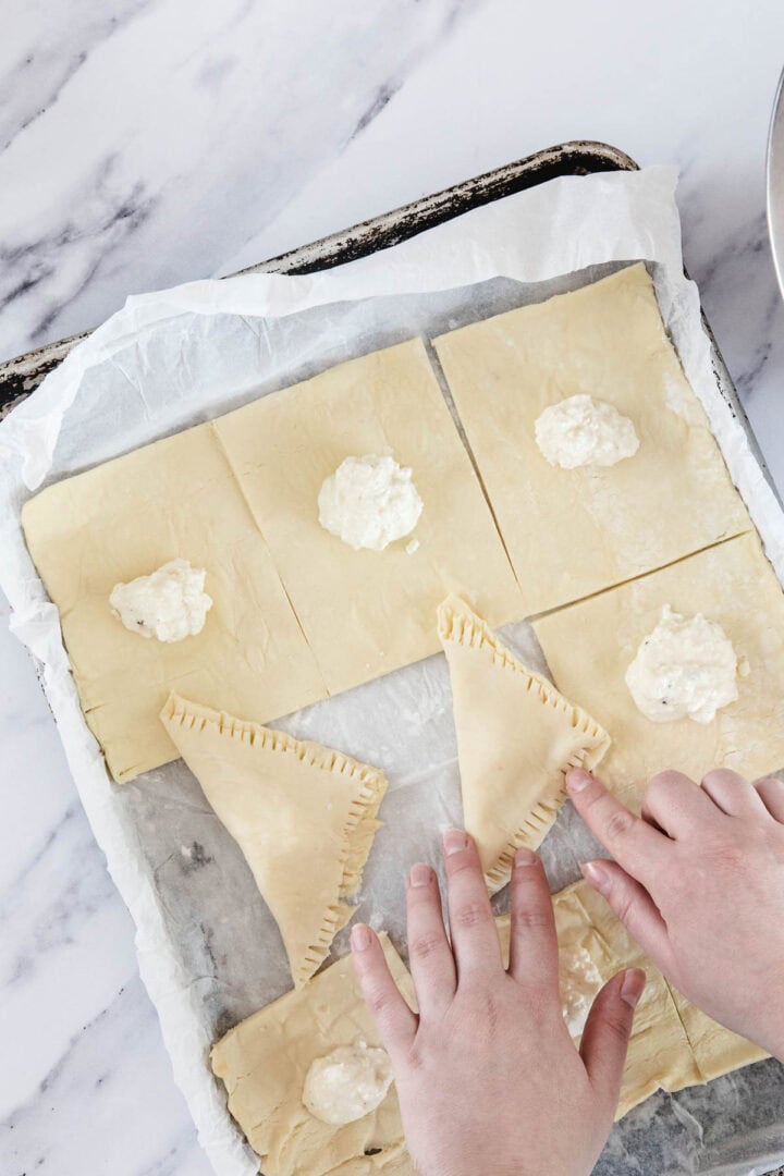 A person's hands folding Cheese Bourekas, puff pastry squares filled with a dollop of cheese mixture, sealing the edges with a fork on a parchment-lined baking sheet. The surface below is white marble.