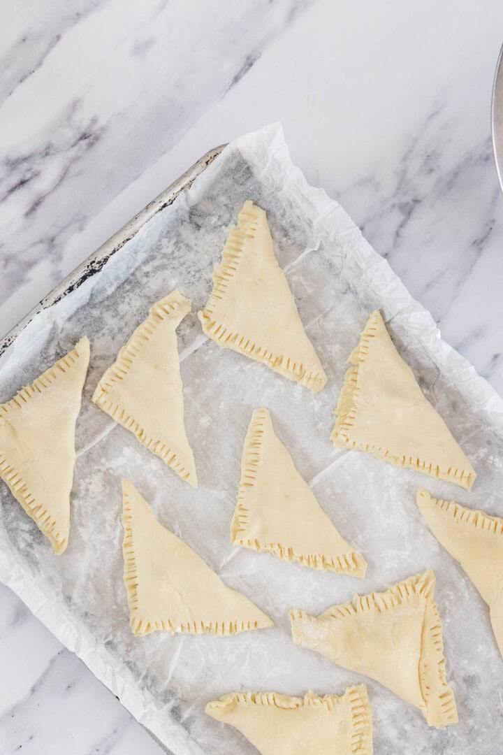 A baking sheet lined with parchment paper holds several unbaked Cheese Bourekas triangles, each with crimped edges, on a marble countertop.