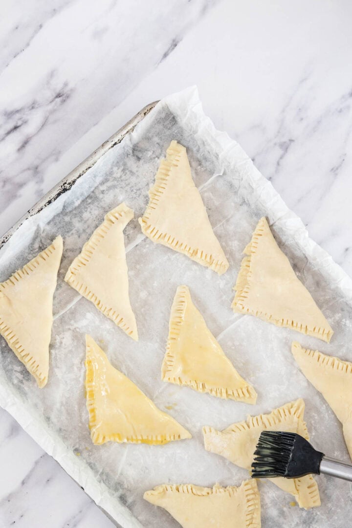 A baking tray lined with parchment paper holds several uncooked, triangular cheese bourekas. A pastry brush is being used to apply an egg wash to one of the turnovers. Marble surface underneath.