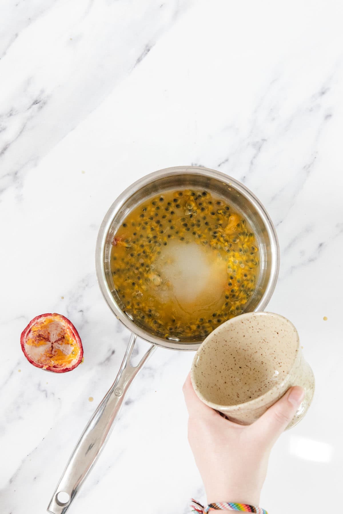 A hand holds a beige cup above a saucepan containing passion fruit pulp and sugar—the perfect base for a Passionfruit Cheesecake. An empty passion fruit shell rests on the white marble countertop beside the saucepan.