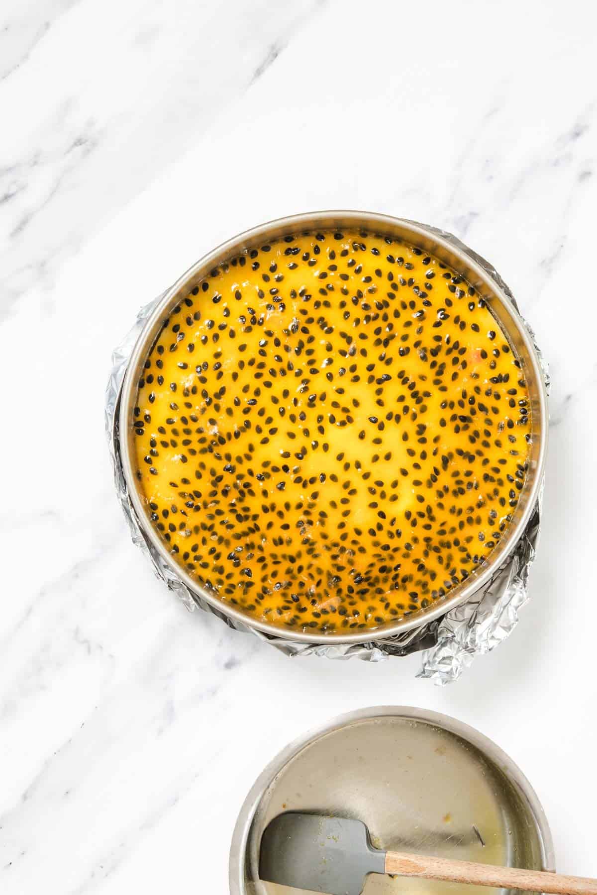 A round Passionfruit Cheesecake covered in yellow glaze with black seeds sits in a metal pan lined with foil on a white marble surface. A spatula and a bowl are visible nearby.