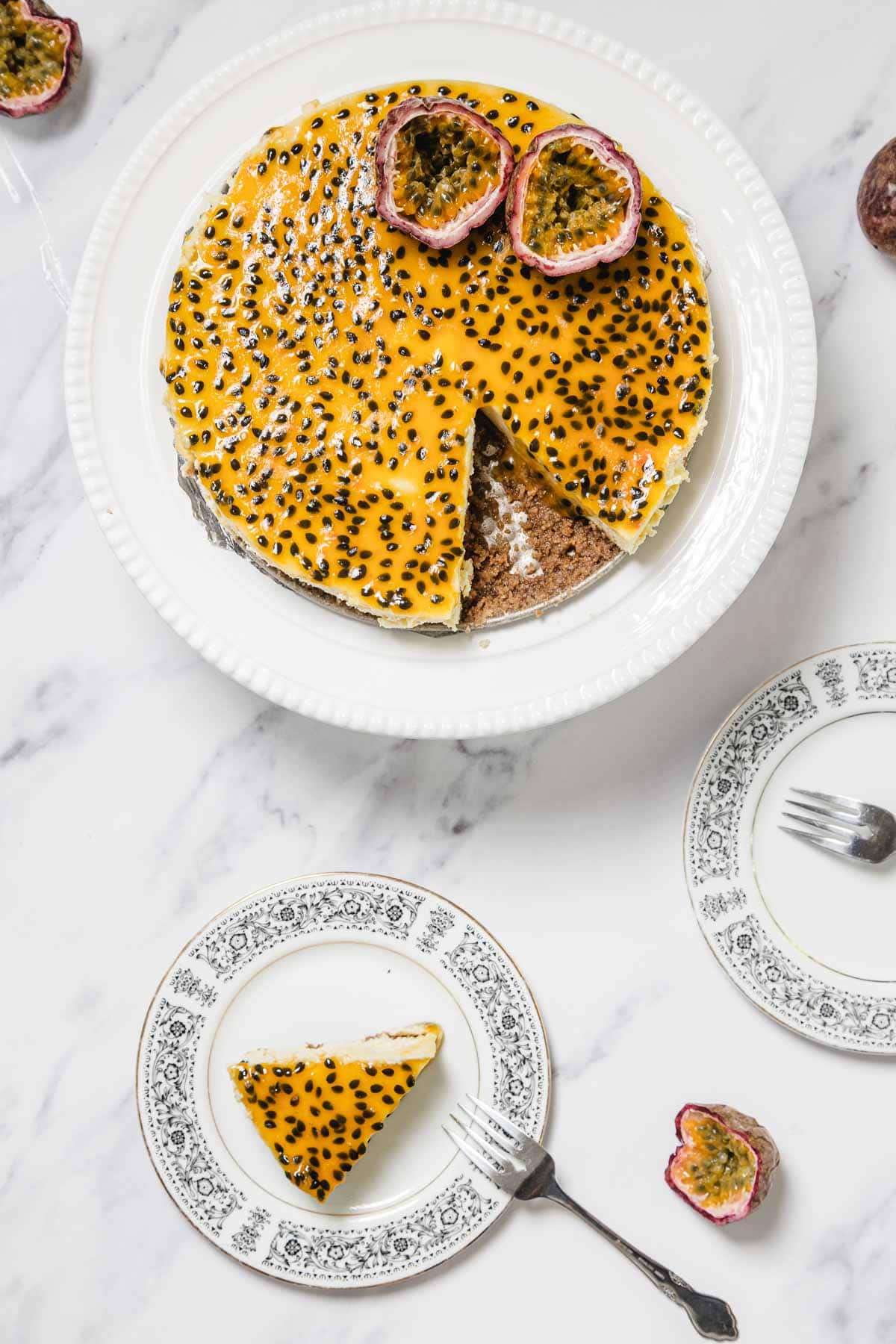 A round Passionfruit Cheesecake with black seeds on top has one slice removed and placed on a small, patterned plate. Halved passion fruits garnish both decorative plates for an extra touch of freshness.