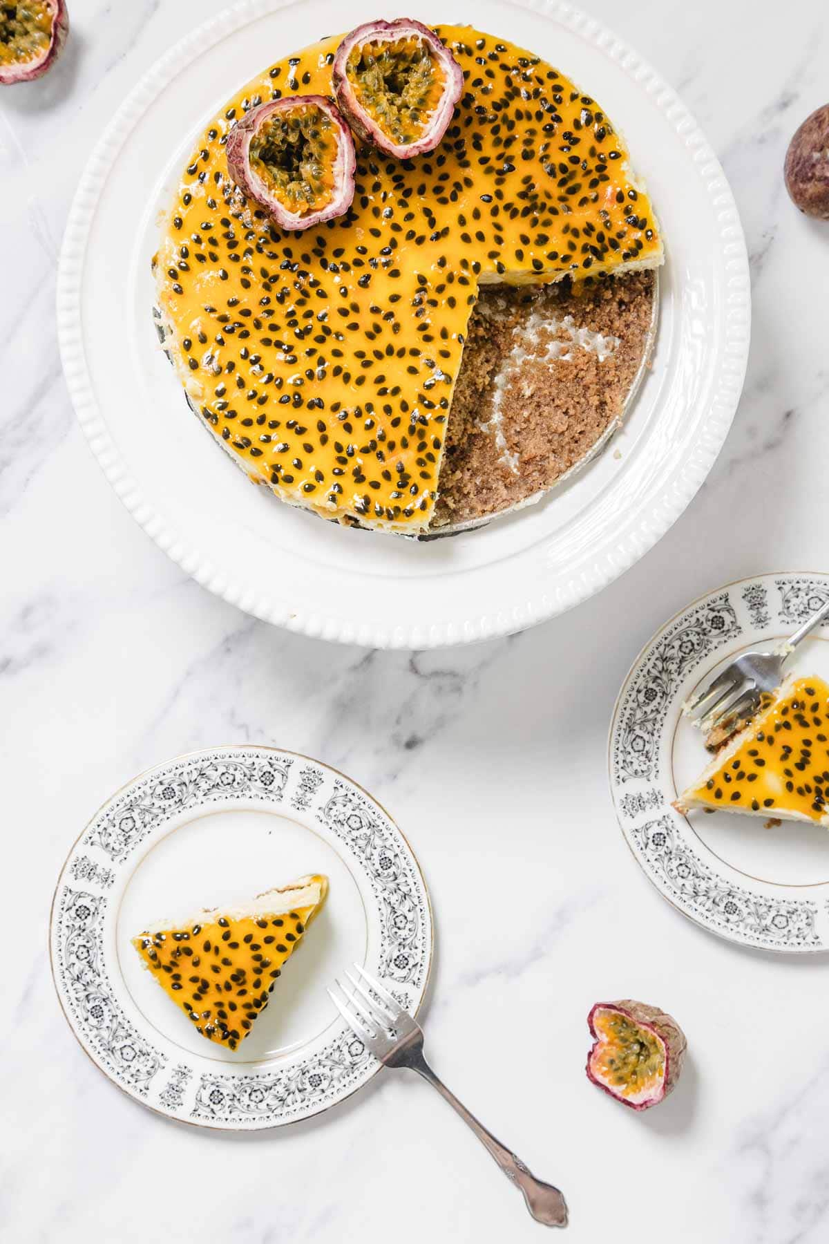 A Passionfruit Cheesecake with a slice removed is displayed on a white plate. Two slices of the cheesecake are served on patterned plates, with halved passionfruits nearby on a white marble surface.
