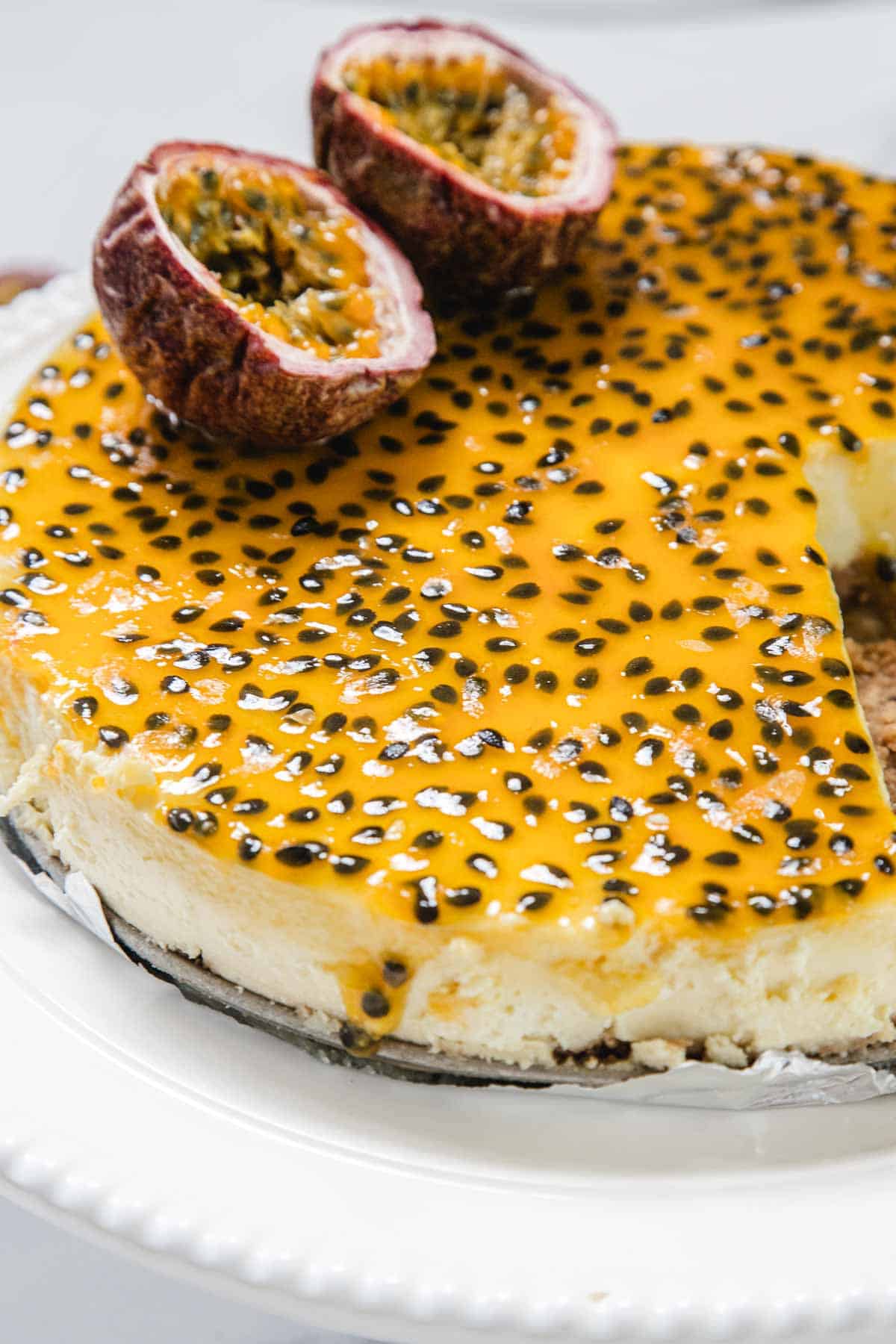 A Passionfruit Cheesecake topped with a glossy passion fruit glaze and black seeds sits on a white plate. Two halved passion fruits garnish the top, while a removed slice reveals the creamy interior of this tropical dessert.