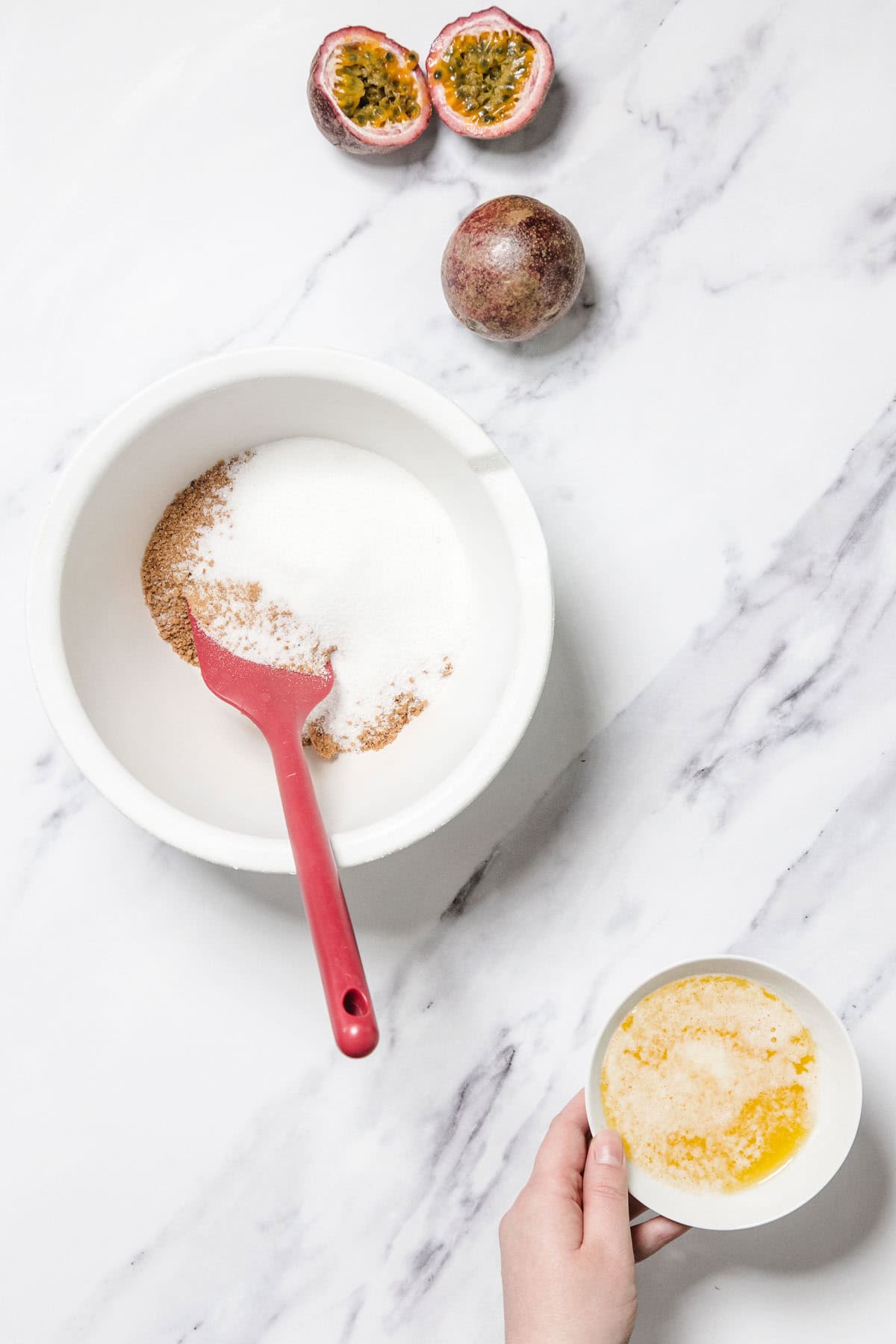 A white bowl with sugar and cinnamon and a red spatula sits on a marble surface. Nearby are two passion fruits, one cut open, a hand holding melted butter, perfect ingredients for making Passionfruit Cheesecake.