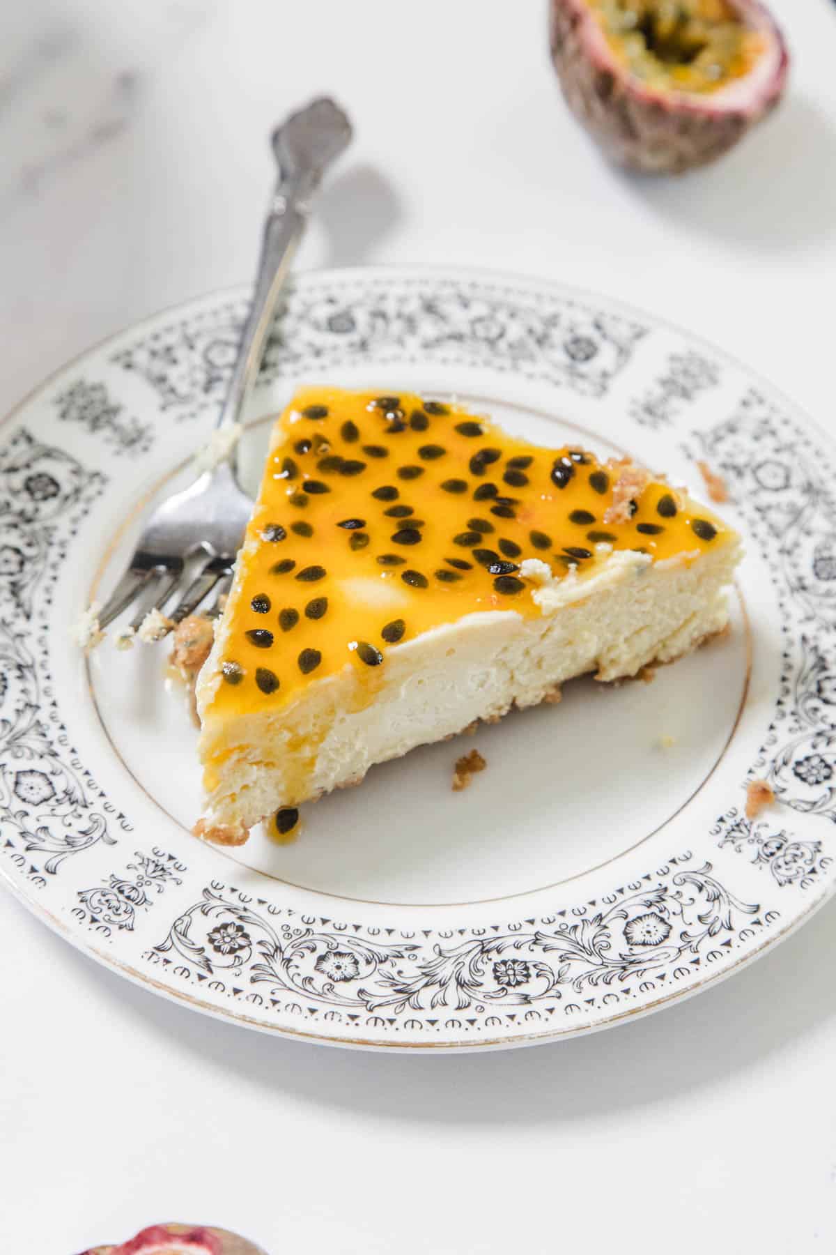 A slice of Passionfruit Cheesecake topped with seeds and sauce sits on a decorative plate with a fork. In the background, a halved passionfruit rests on a white surface, highlighting the dessert’s tropical flair.