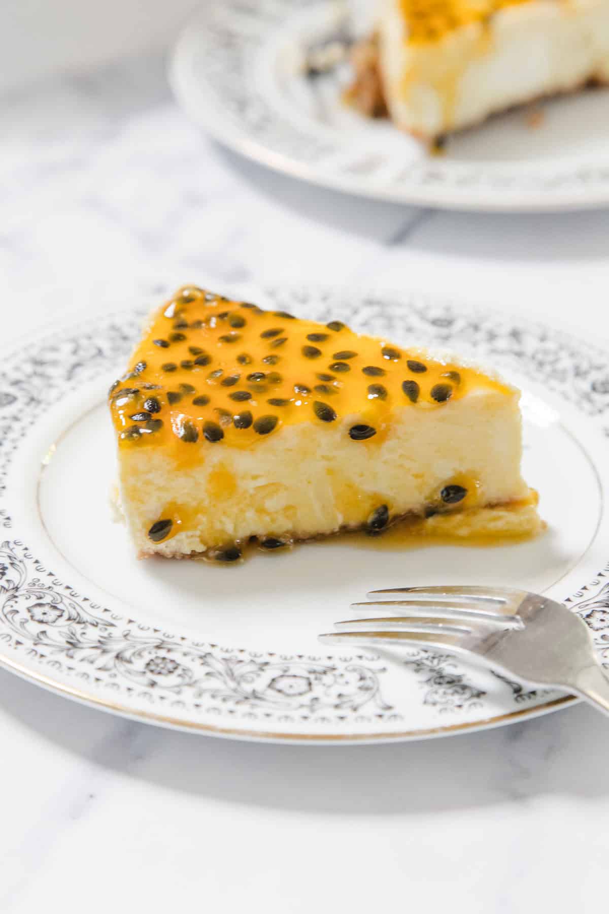 A slice of Passionfruit Cheesecake topped with tangy passionfruit pulp sits on a decorative white plate with a floral pattern. A fork rests beside the cheesecake slice, and another plate appears in the background.