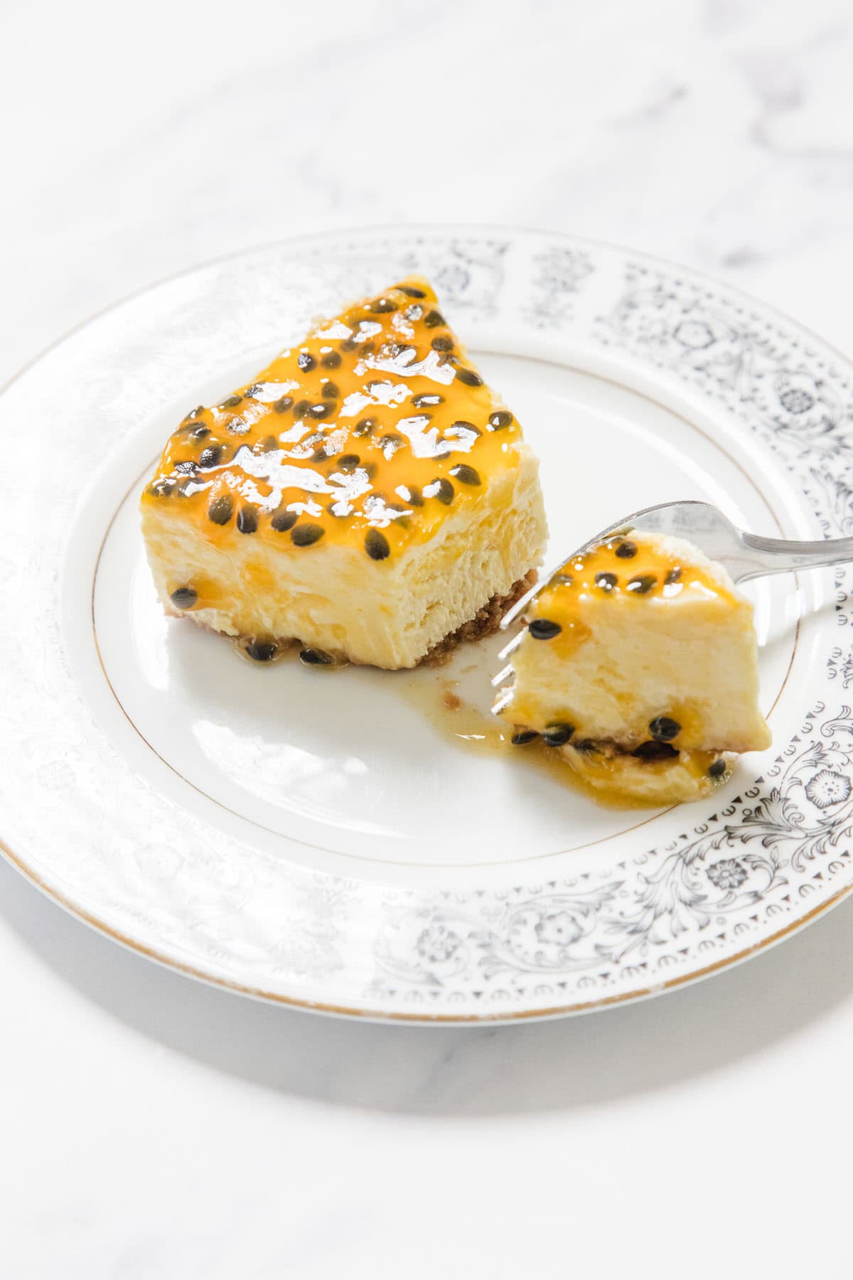 A slice of Passionfruit Cheesecake topped with tangy passion fruit pulp sits on a decorative white plate, with a fork cutting into the creamy dessert.
