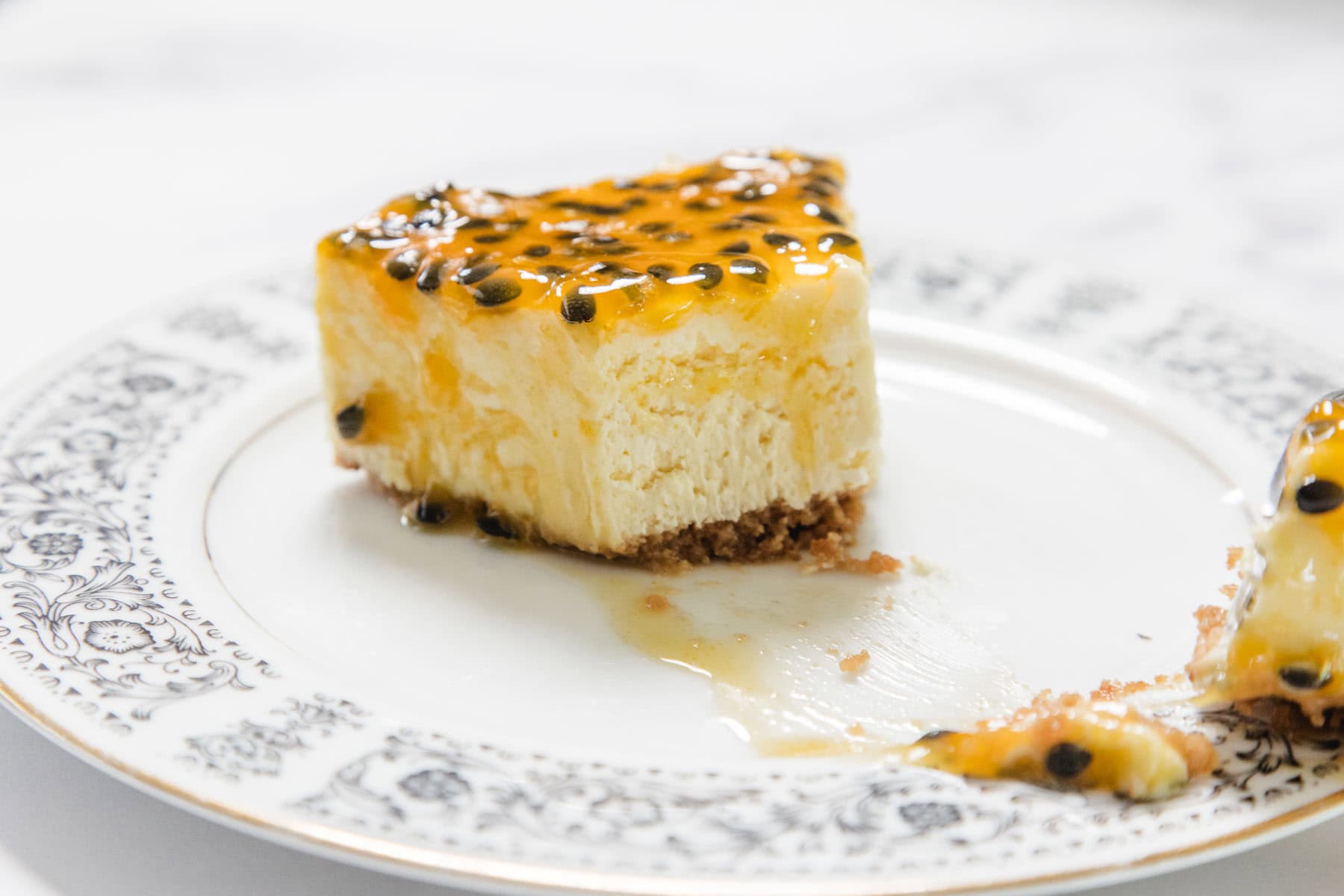 A slice of Passionfruit Cheesecake with a crumbly base and tangy topping sits on a decorative white plate with a floral border. Some crumbs and a small piece of cake rest beside the main slice.